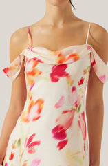 A woman is wearing a sleeveless, off-the-shoulder dress with thin spaghetti straps. The dress has a soft, flowing fabric with a floral pattern featuring large, blurred flowers in shades of pink, red, orange, and green on a light, creamy background. The neckline drapes loosely across the shoulders, creating a relaxed and elegant look.