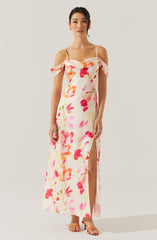 A woman is wearing a long, sleeveless, off-the-shoulder dress with spaghetti straps. The dress features a watercolor floral pattern in shades of pink, red, orange, and green on a white background. It has a slit on one side, revealing her leg. She is also wearing open-toe heels.