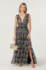 A woman is wearing a sleeveless, maxi-length dress with a deep V neckline. The dress has a black background with a colorful floral pattern including purple, yellow, and green flowers. It features a cinched waist and a high slit on one side, revealing her leg. She is accessorizing with large, round pearl earrings and is carrying a small, light-colored handbag. She is also wearing strappy, open-toe heels.