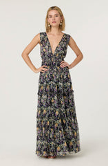 The dress features a sleeveless design with wide shoulder straps and a deep V-neckline. It has a fitted waist and a long, flowing skirt divided into three tiers, each adorned with a vibrant floral pattern. The fabric appears lightweight and semi-sheer, suited for warm weather.