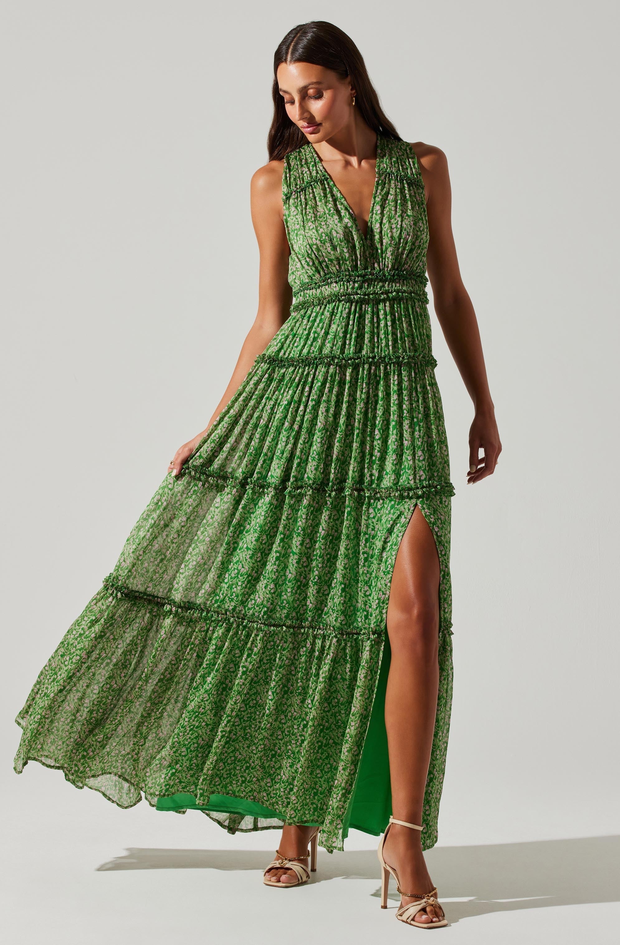 A woman is wearing a sleeveless, green, tiered maxi dress with a deep V neckline. The dress features a small slit on the right side, revealing her leg. The fabric has a floral pattern, and the dress has a gathered waistband. She is wearing beige high-heeled sandals.