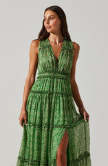 A woman is wearing a sleeveless, green dress with a floral pattern. The dress has a deep V-neckline, an elastic waistband, and tiered ruffles throughout the length. The fabric is sheer in some sections, revealing a slit that shows her leg.