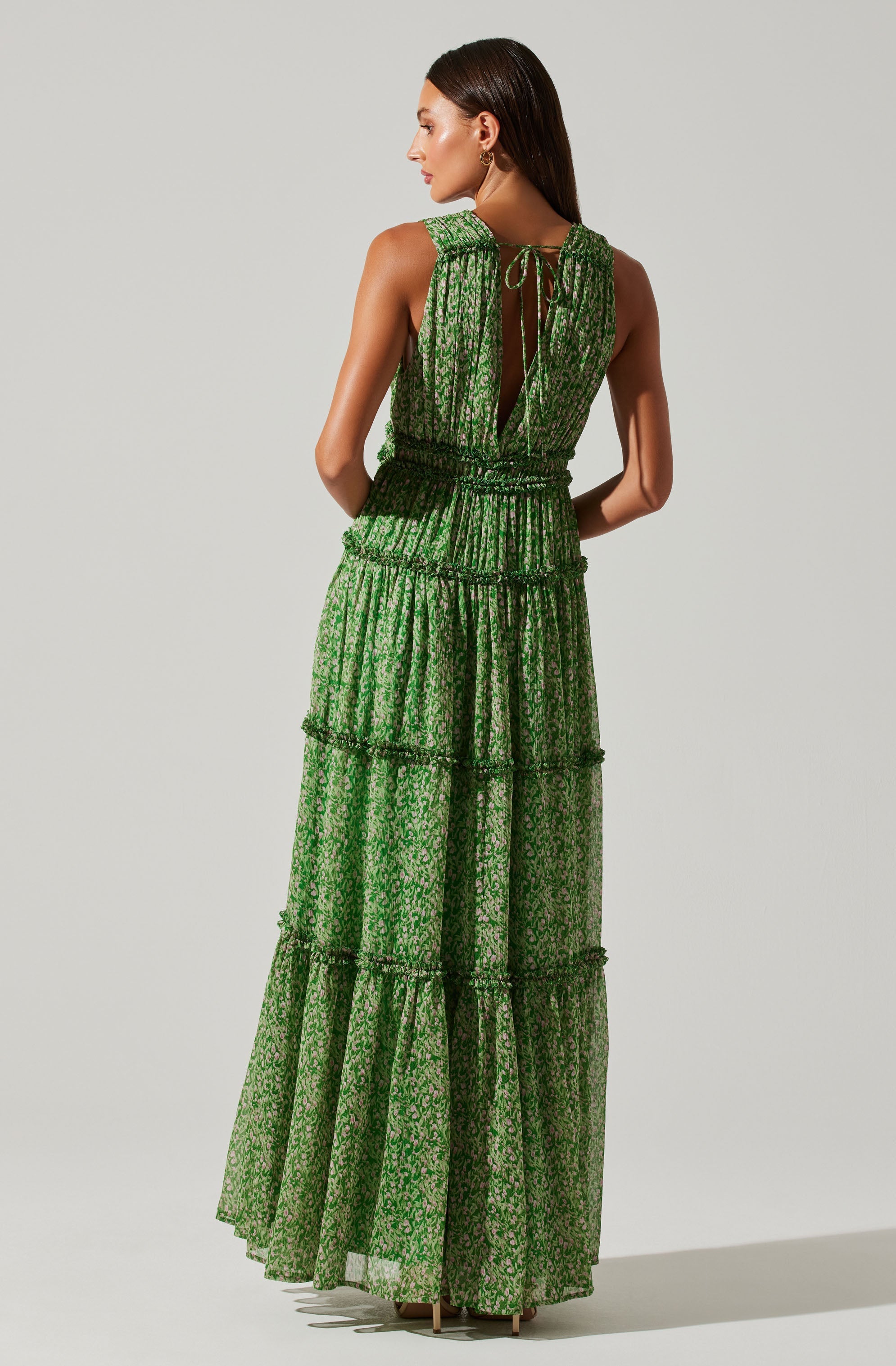 A woman is wearing a sleeveless, floor-length dress made of sheer, green fabric with a floral pattern. The dress features multiple horizontal tiers and a back opening with a tie closure at the neckline. The fabric appears lightweight and flowy.