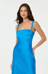 A woman is wearing a sleeveless, bright blue satin dress with spaghetti straps. The dress has a fitted bodice with a subtle seam detail across the chest and a smooth, flowing skirt. She is accessorized with large, round, shiny earrings.
