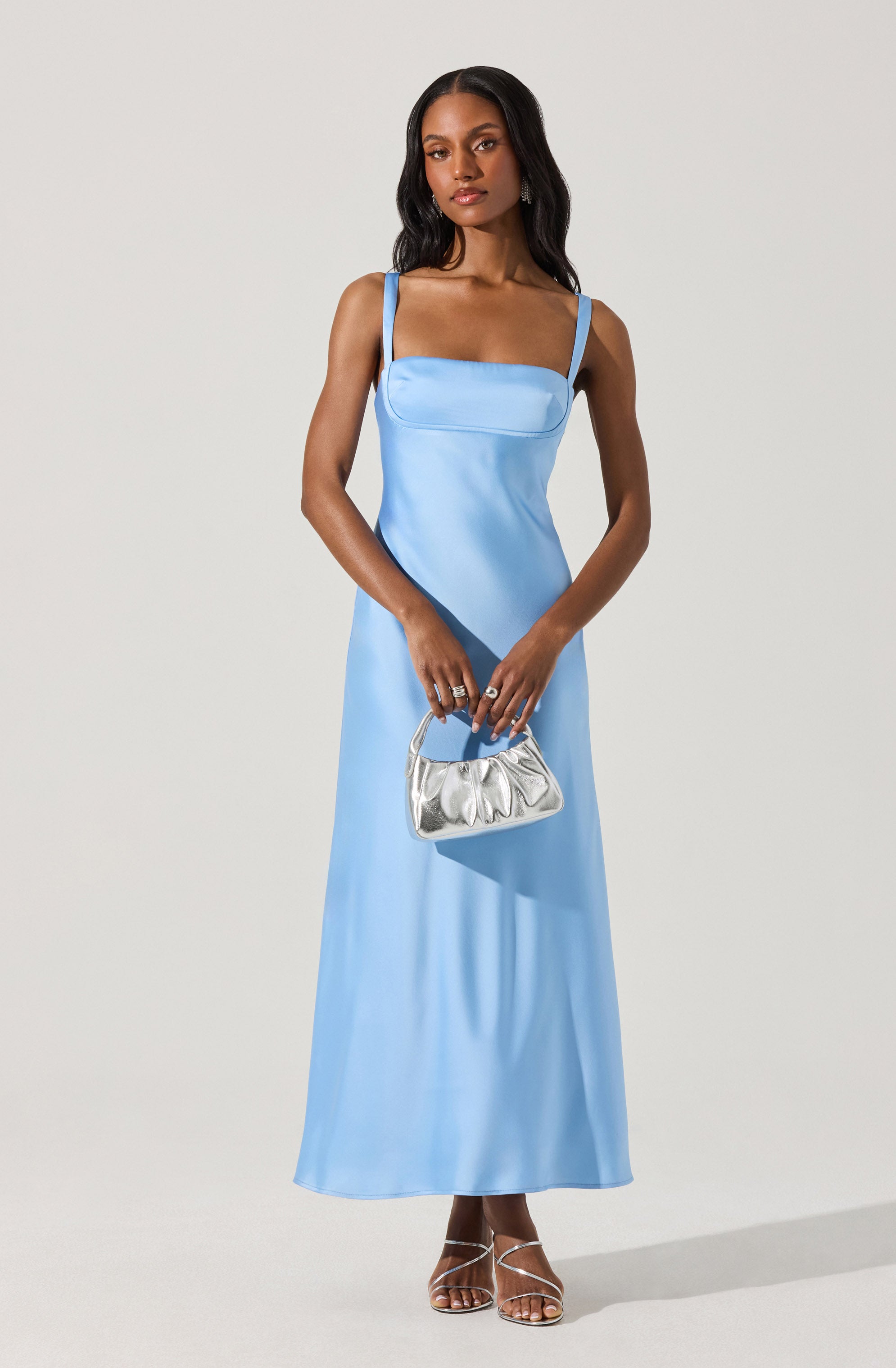 A woman is wearing a light blue, sleeveless, floor-length dress with a fitted bodice and thin shoulder straps. She is holding a small, metallic silver handbag and is wearing silver strappy high-heeled sandals.