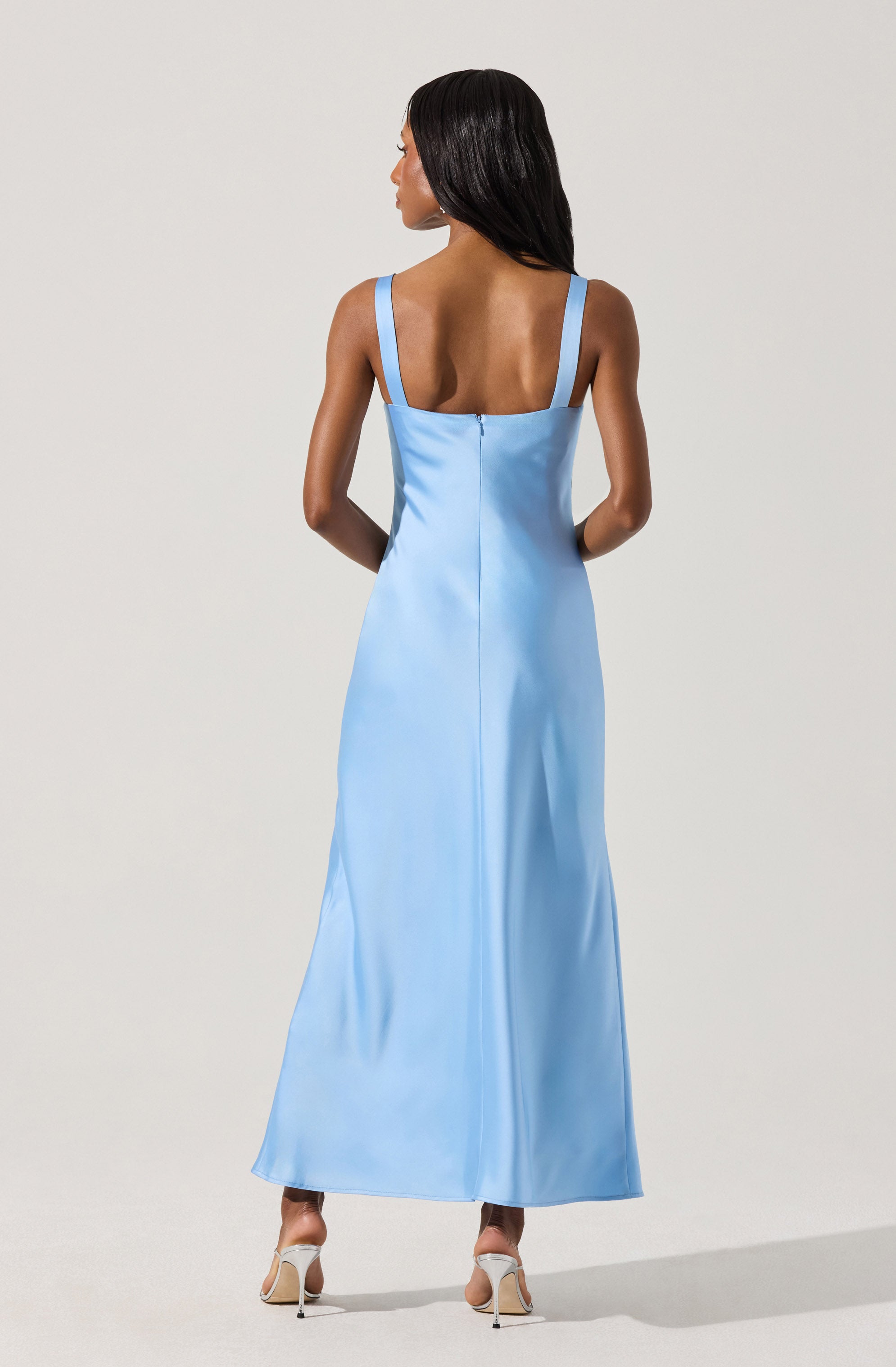 A woman is wearing a long, light blue satin evening dress with wide shoulder straps. The dress has a back zipper and a full-length, flowing skirt. She is also wearing beige high-heeled shoes.