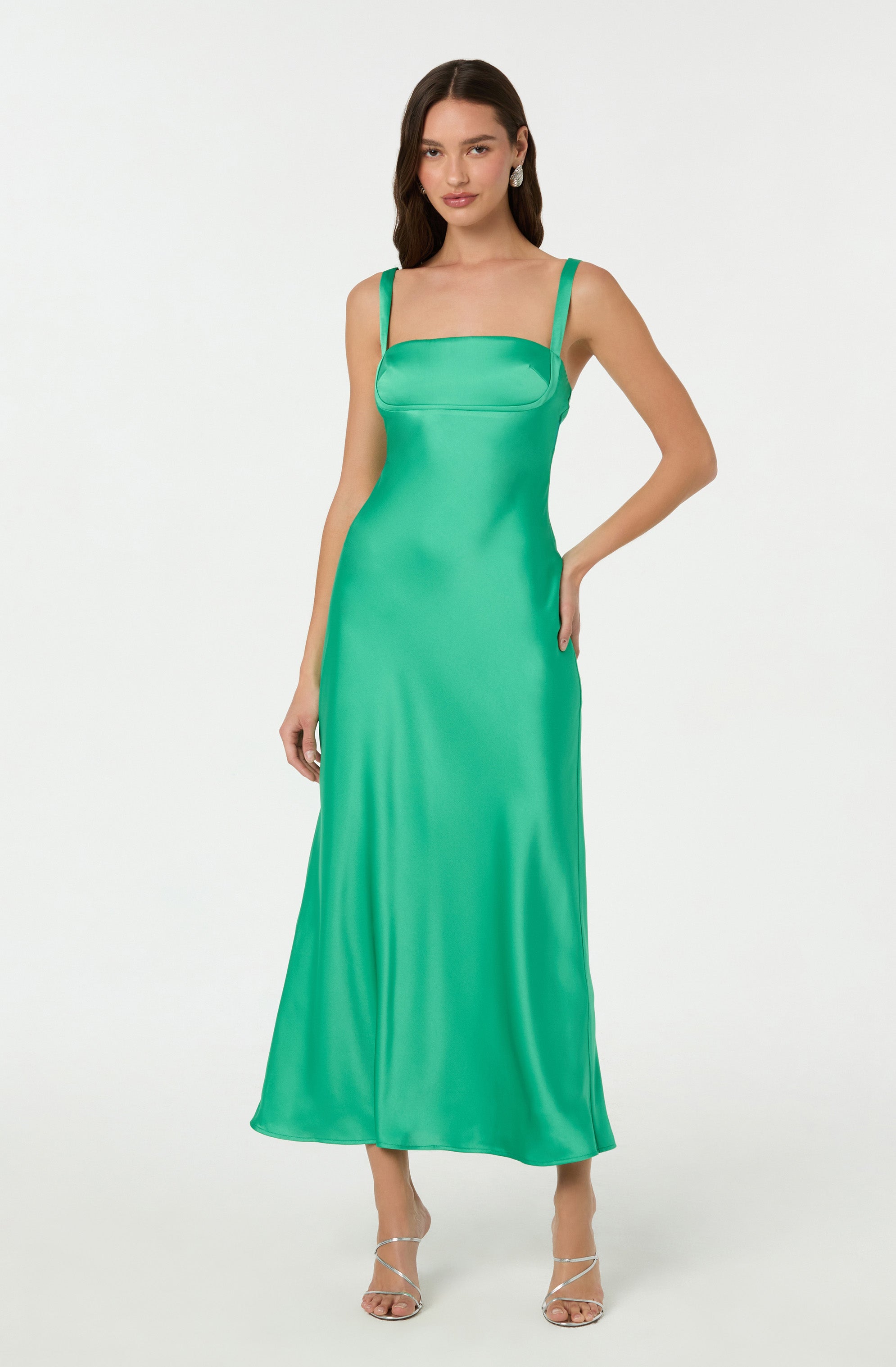 The dress is a sleeveless, full-length gown made from a silky, satin-like fabric with a shiny finish. It features a fitted bodice with a straight neckline and thin shoulder straps. The dress is a vibrant shade of green and has a smooth, flowing silhouette that falls gracefully to the floor. The woman is also wearing open-toe, strappy high-heeled sandals.