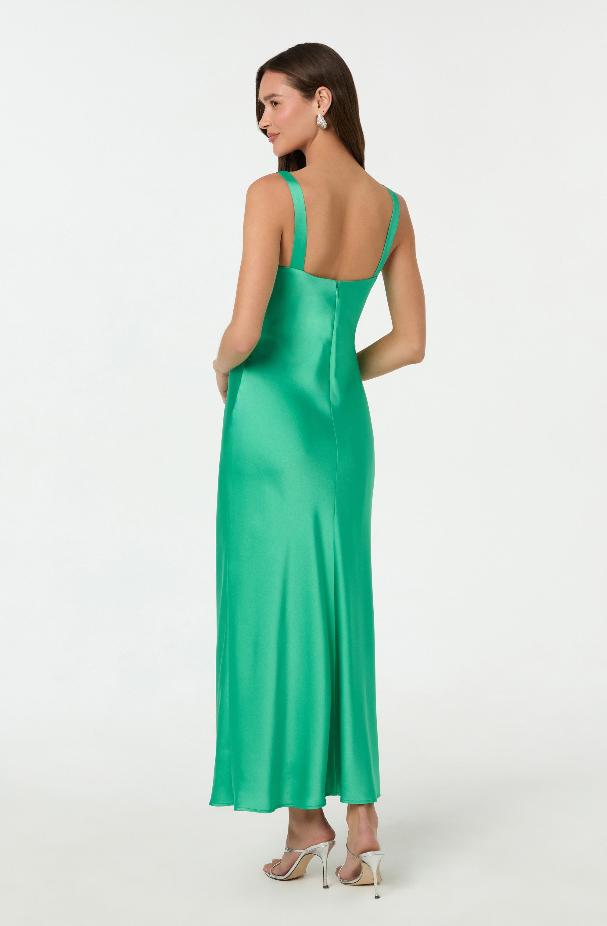 A woman is wearing a floor-length, sleeveless gown in a vibrant, shiny green satin fabric. The dress has wide shoulder straps and a fitted bodice, flowing smoothly down to the hem. She is also wearing white high-heeled shoes with thin straps and large, sparkling earrings.