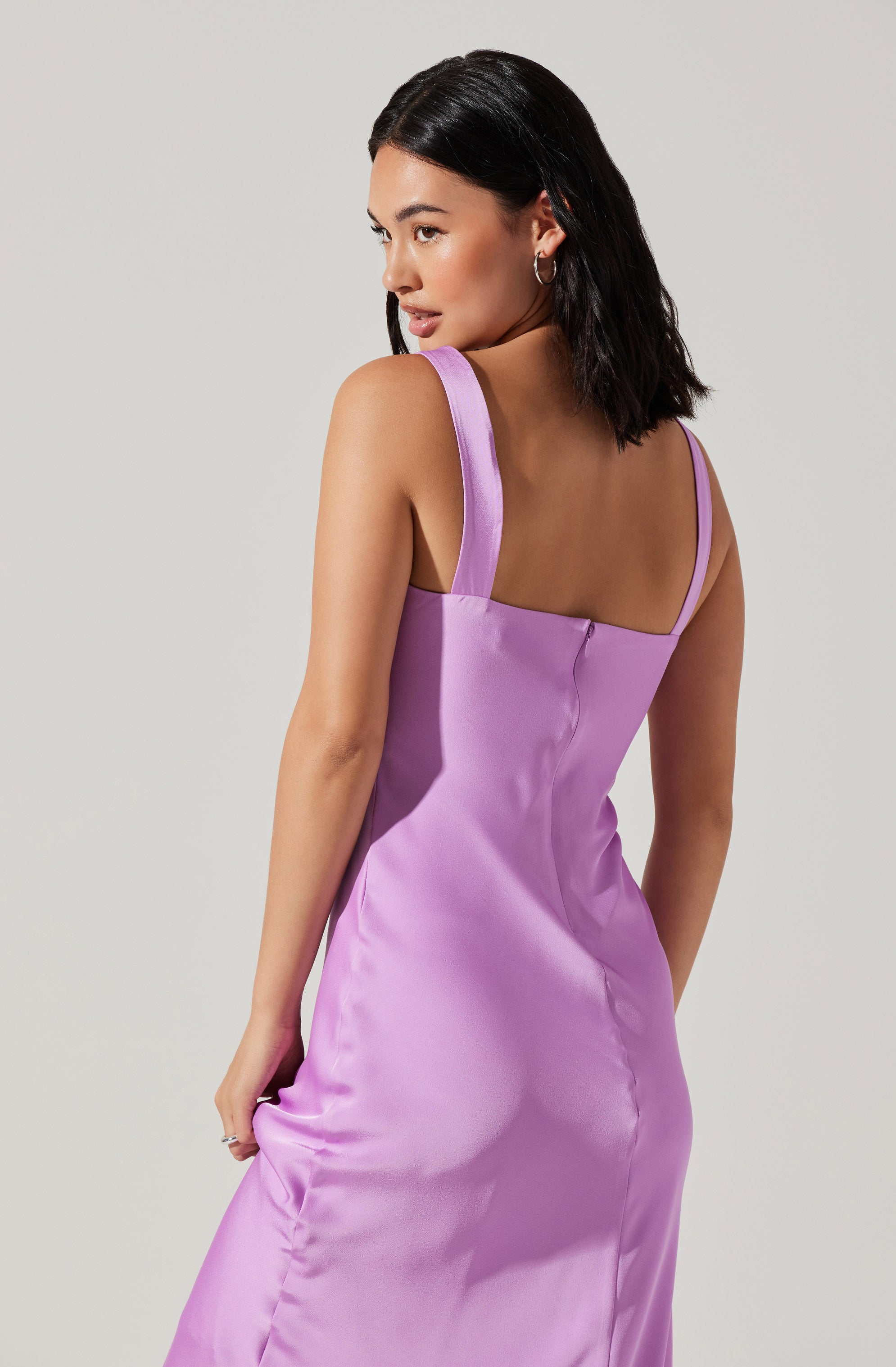 A woman is wearing a sleeveless, lavender satin dress with wide shoulder straps. The dress has a fitted bodice and a smooth, shiny fabric that drapes down to the floor. She has short, dark hair and is accessorized with hoop earrings.