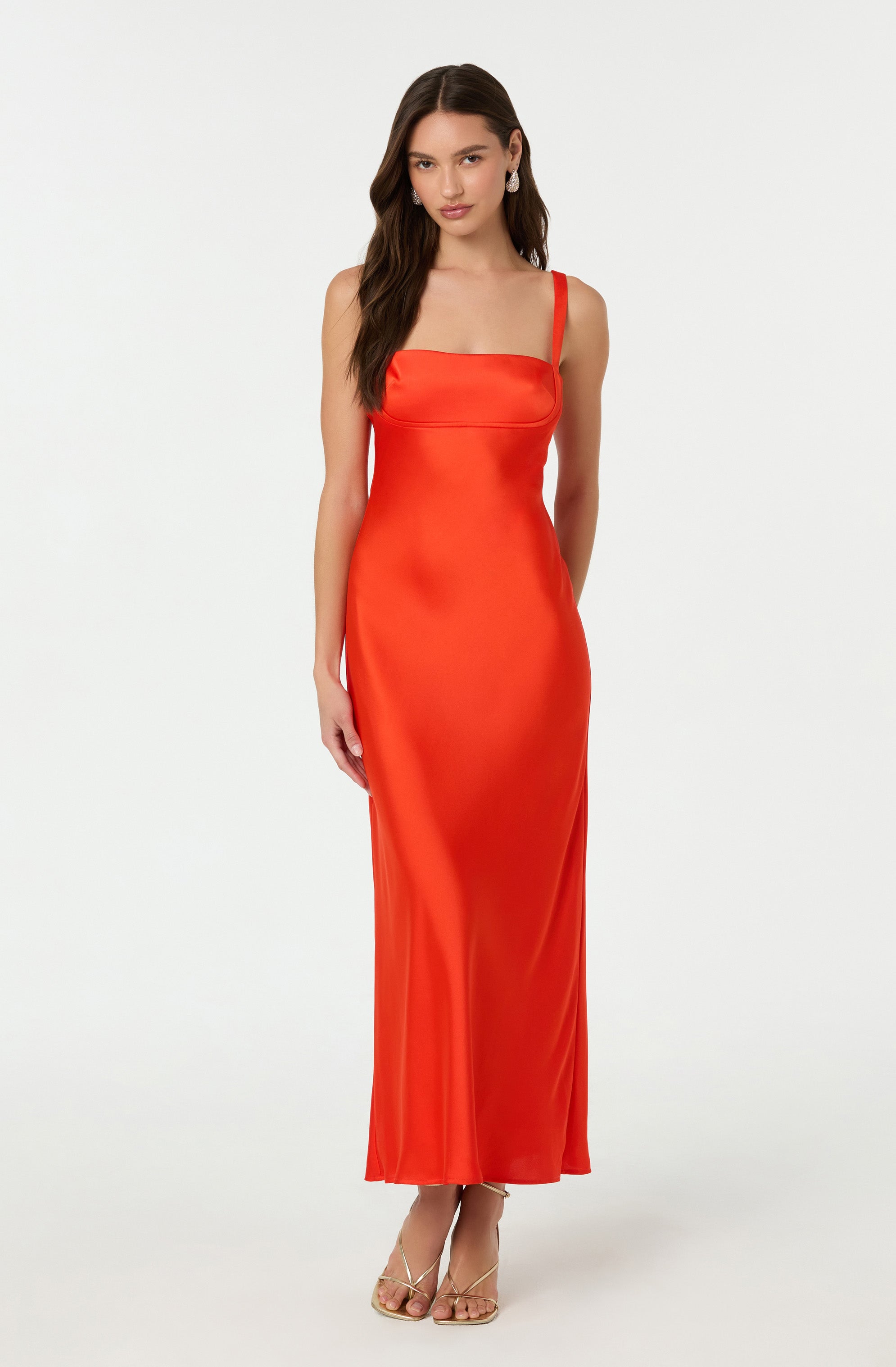 A woman is wearing a sleeveless, floor-length satin dress in a vibrant red-orange color. The dress features a fitted bodice with a square neckline and narrow shoulder straps. She is also wearing nude strappy high-heeled sandals and has long dark hair styled in loose waves.