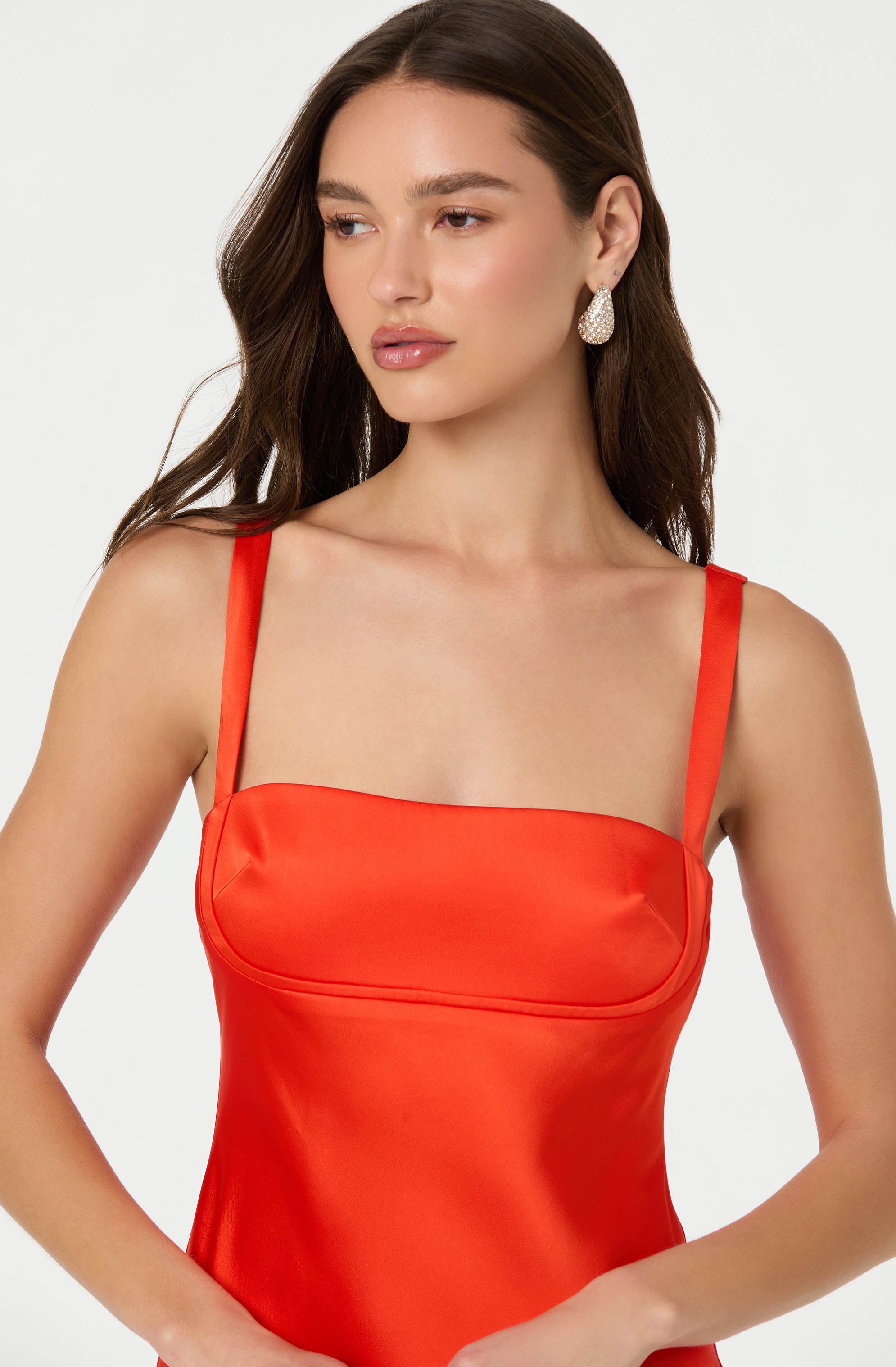 A woman is wearing a bright red satin dress with thin shoulder straps. The dress has a fitted bodice with a square neckline. She is accessorized with large, teardrop-shaped earrings.