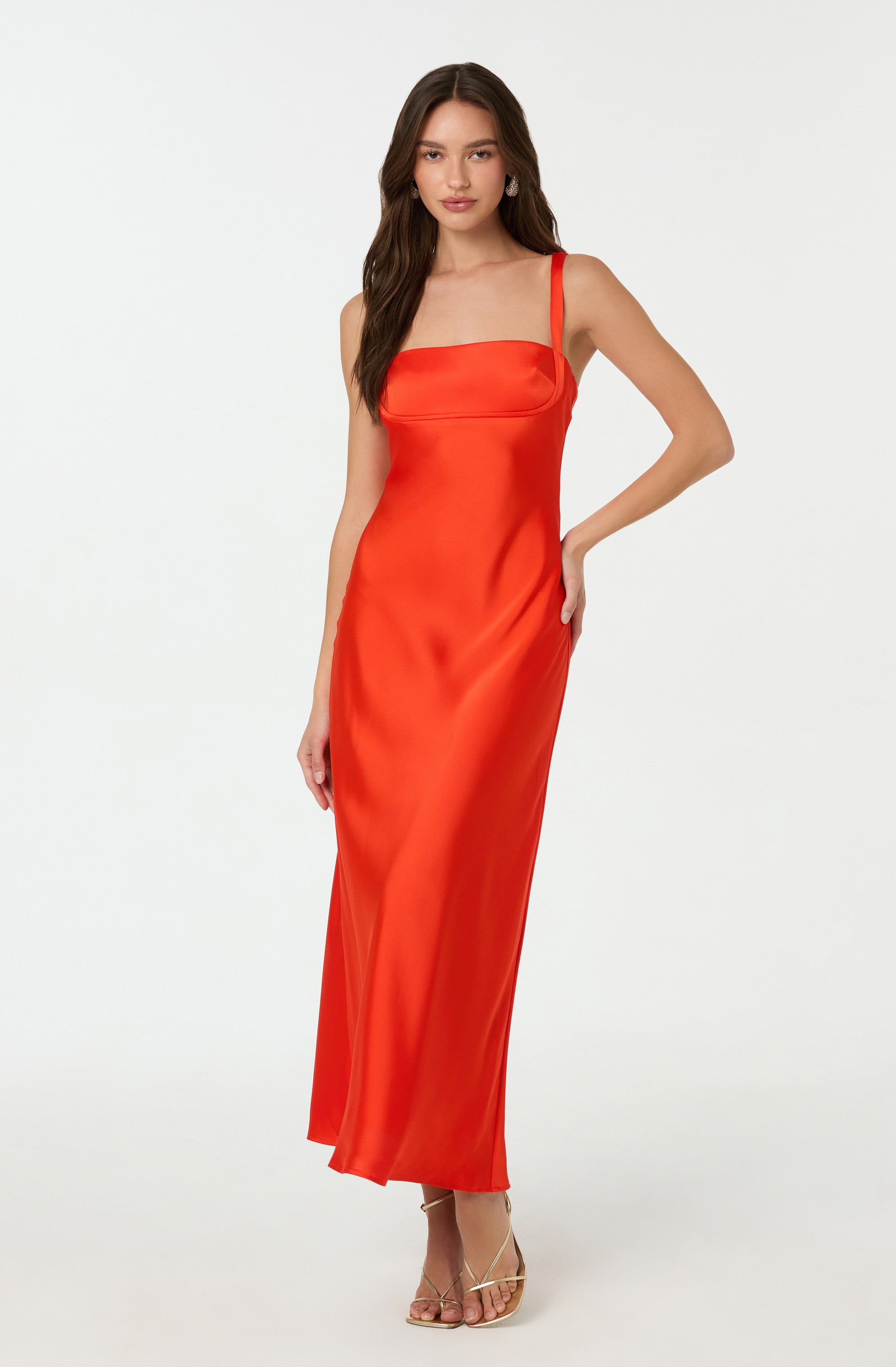 A woman is wearing a long, sleeveless, coral-red dress with wide shoulder straps and a fitted bodice with a subtle horizontal seam detail across the chest. The dress has a smooth, shiny satin-like fabric that flows down to the floor. She pairs the dress with nude, strappy high-heeled sandals.