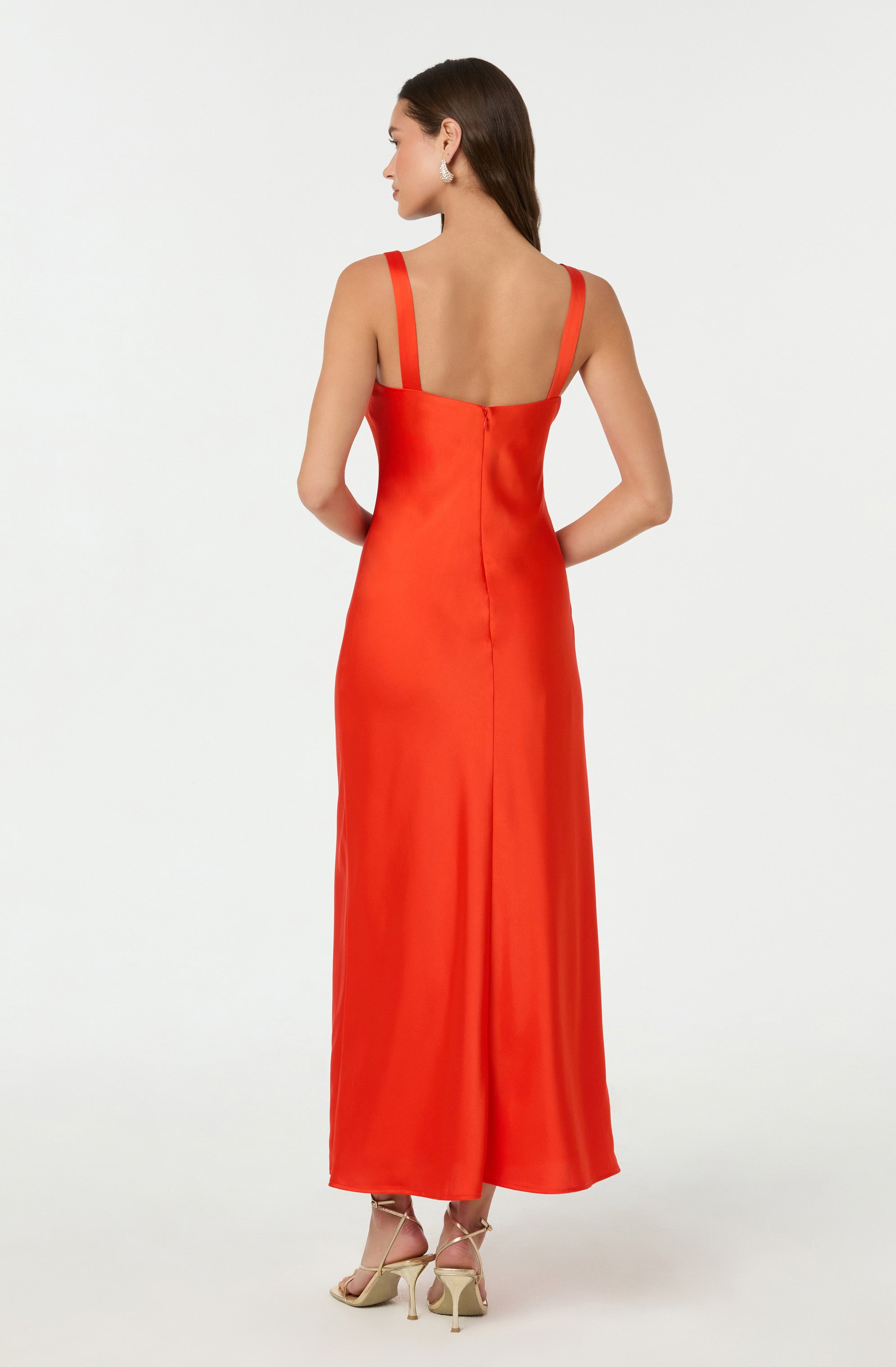 A woman is wearing a long, sleeveless, red satin dress with wide straps. The dress has a fitted bodice and a straight, flowing skirt that reaches her ankles. She is also wearing beige high-heeled shoes.