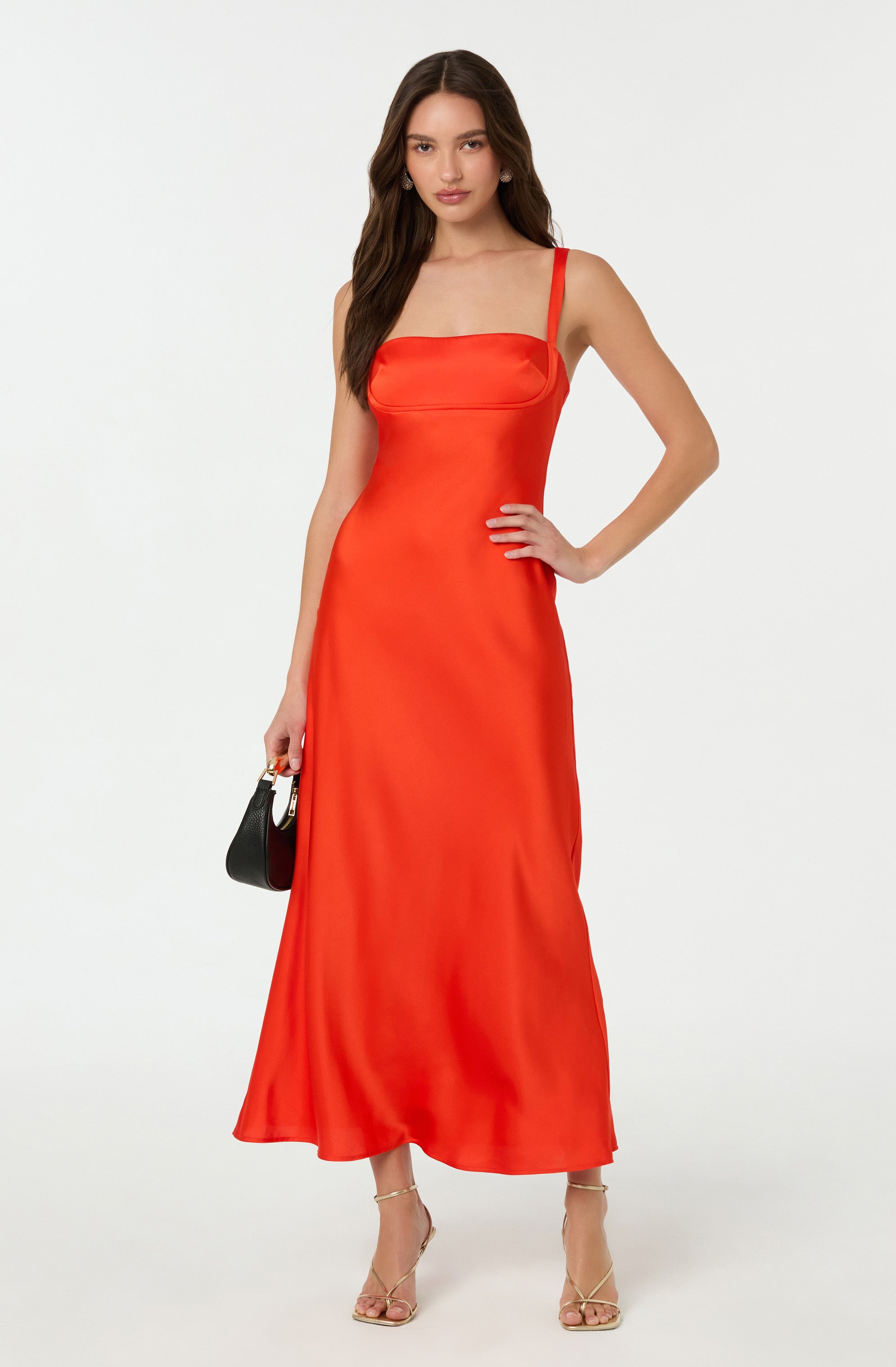 A woman is wearing a long, flowing red satin dress with a fitted bodice and wide shoulder straps. She is holding a small black handbag in her right hand and is wearing beige high-heeled sandals.