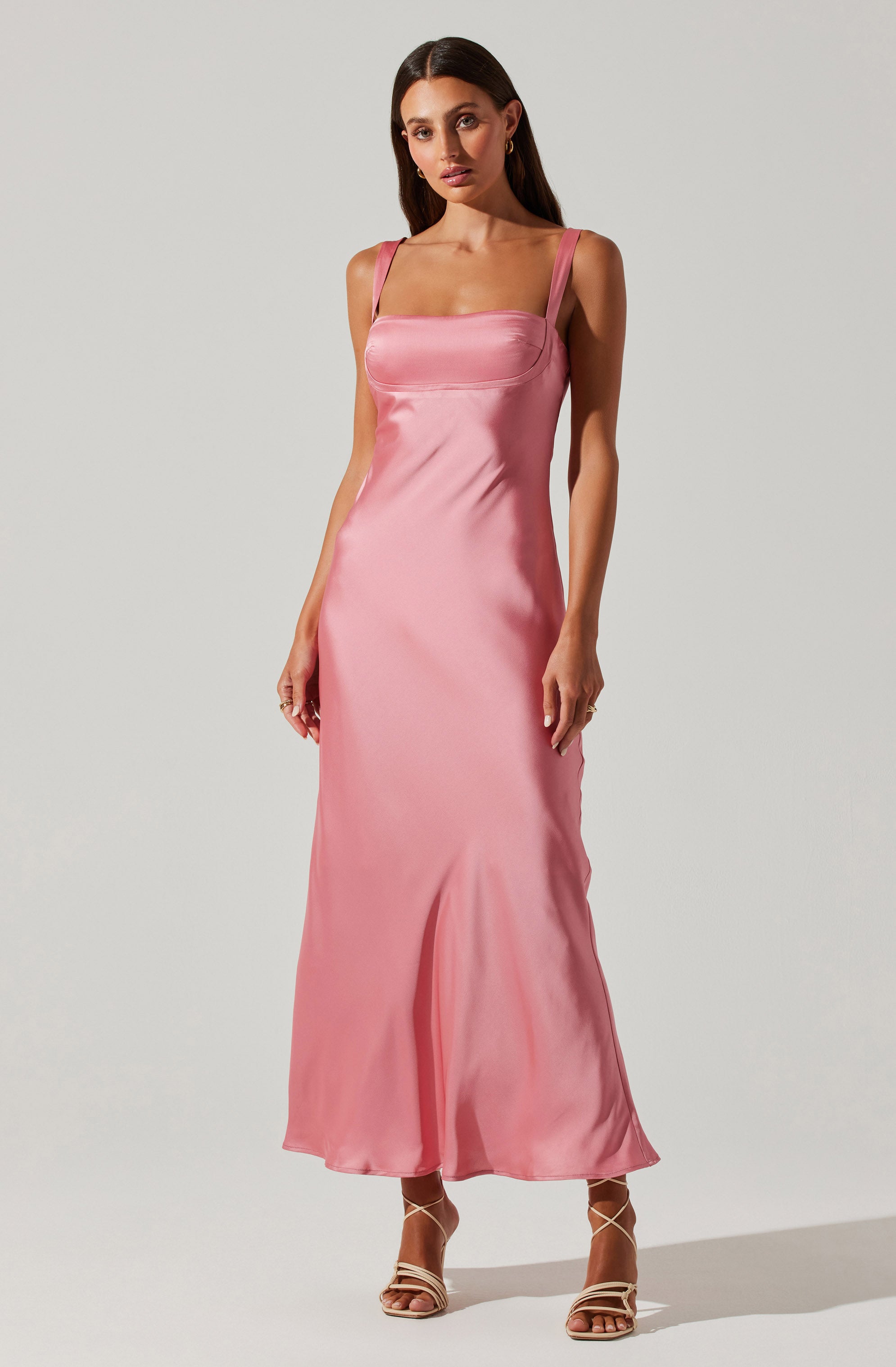 A woman is wearing a long, silky pink satin dress with wide shoulder straps. The dress has a fitted bodice with a horizontal seam detail just below the bust and flows down to the floor with a slight flare at the hem. She completes the look with strappy high-heeled sandals.