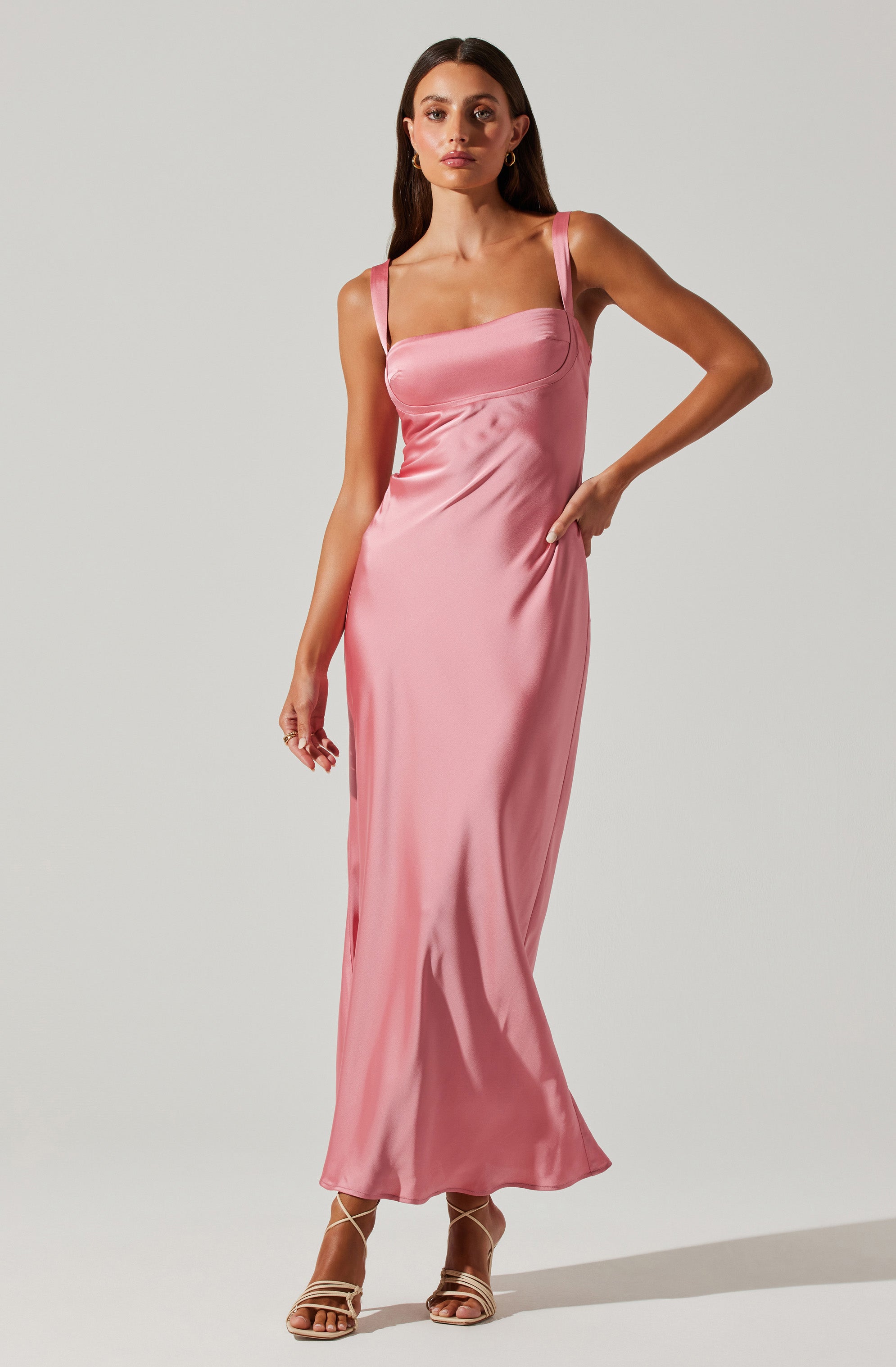 A woman is wearing a long, silky pink dress with wide shoulder straps. The dress has a fitted bodice and flows down to the floor, creating an elegant silhouette. She is also wearing open-toe high-heeled sandals.