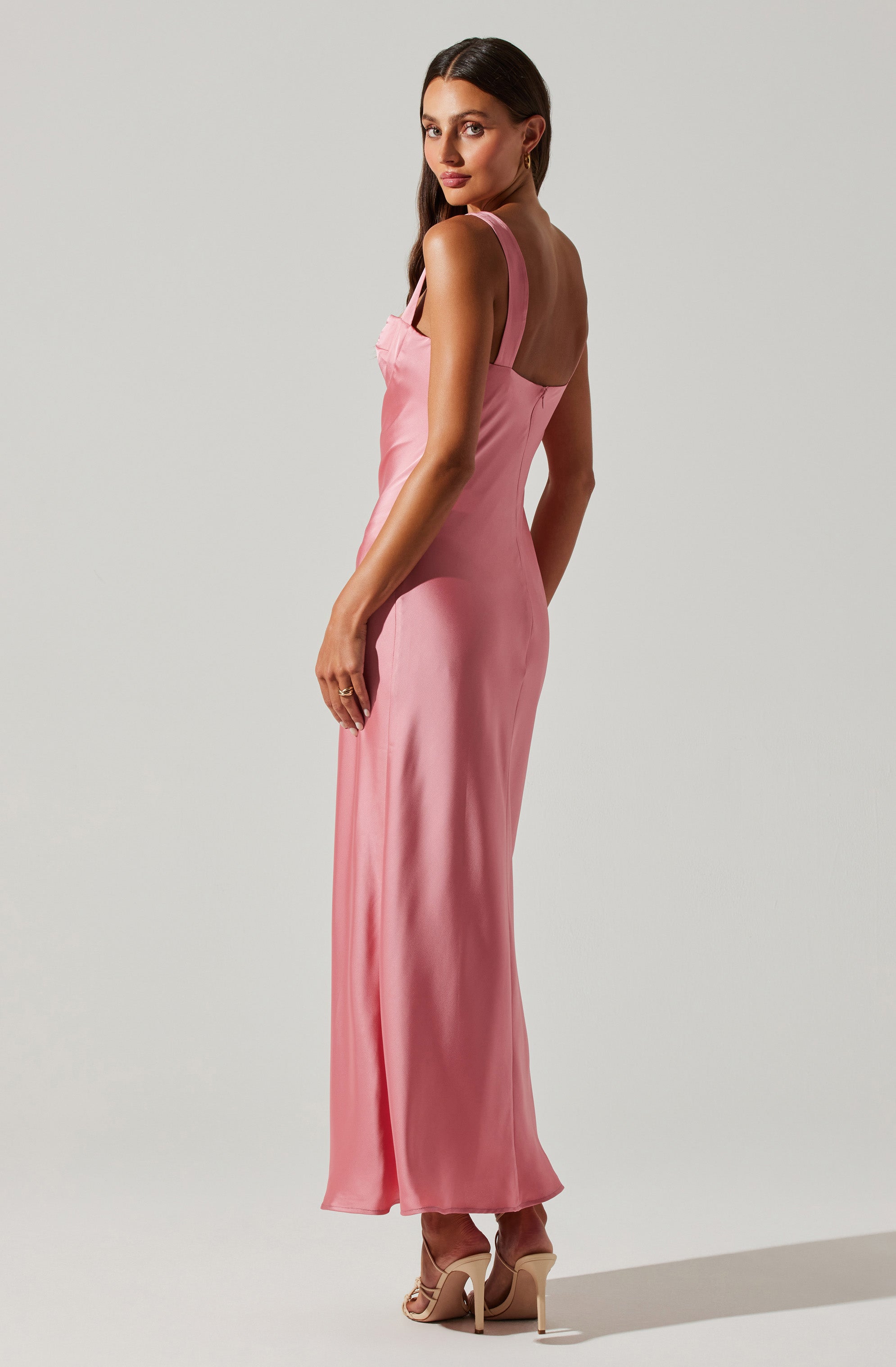 A woman is wearing a sleeveless, light pink, floor-length satin dress with wide shoulder straps. The dress has a fitted bodice and a flowing skirt. She is also wearing nude high-heeled sandals.