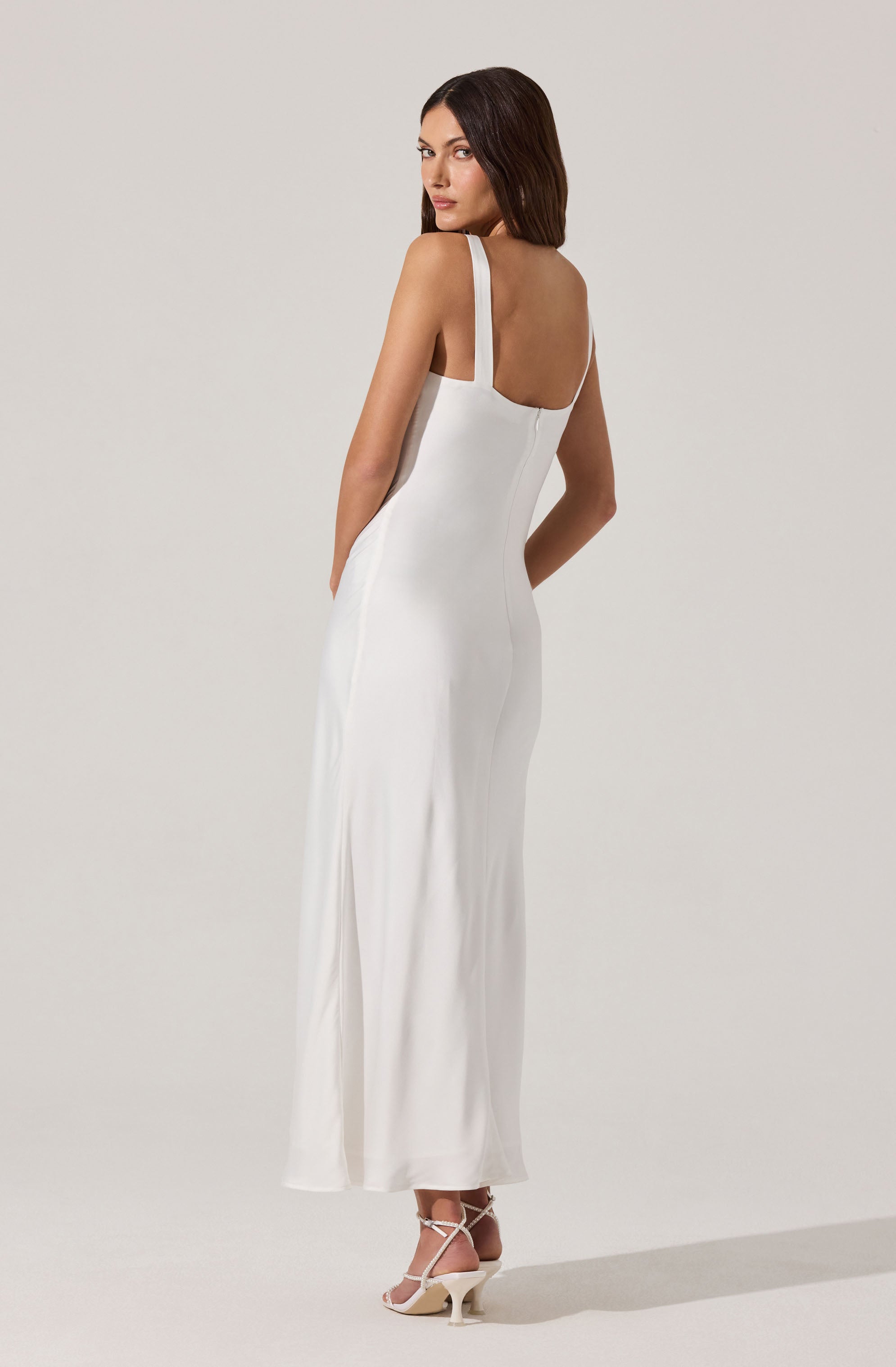 A woman is wearing a long, elegant white dress with thin shoulder straps. The dress has a fitted bodice and flows down to the floor, creating a sleek silhouette. She is also wearing strappy high-heeled shoes.