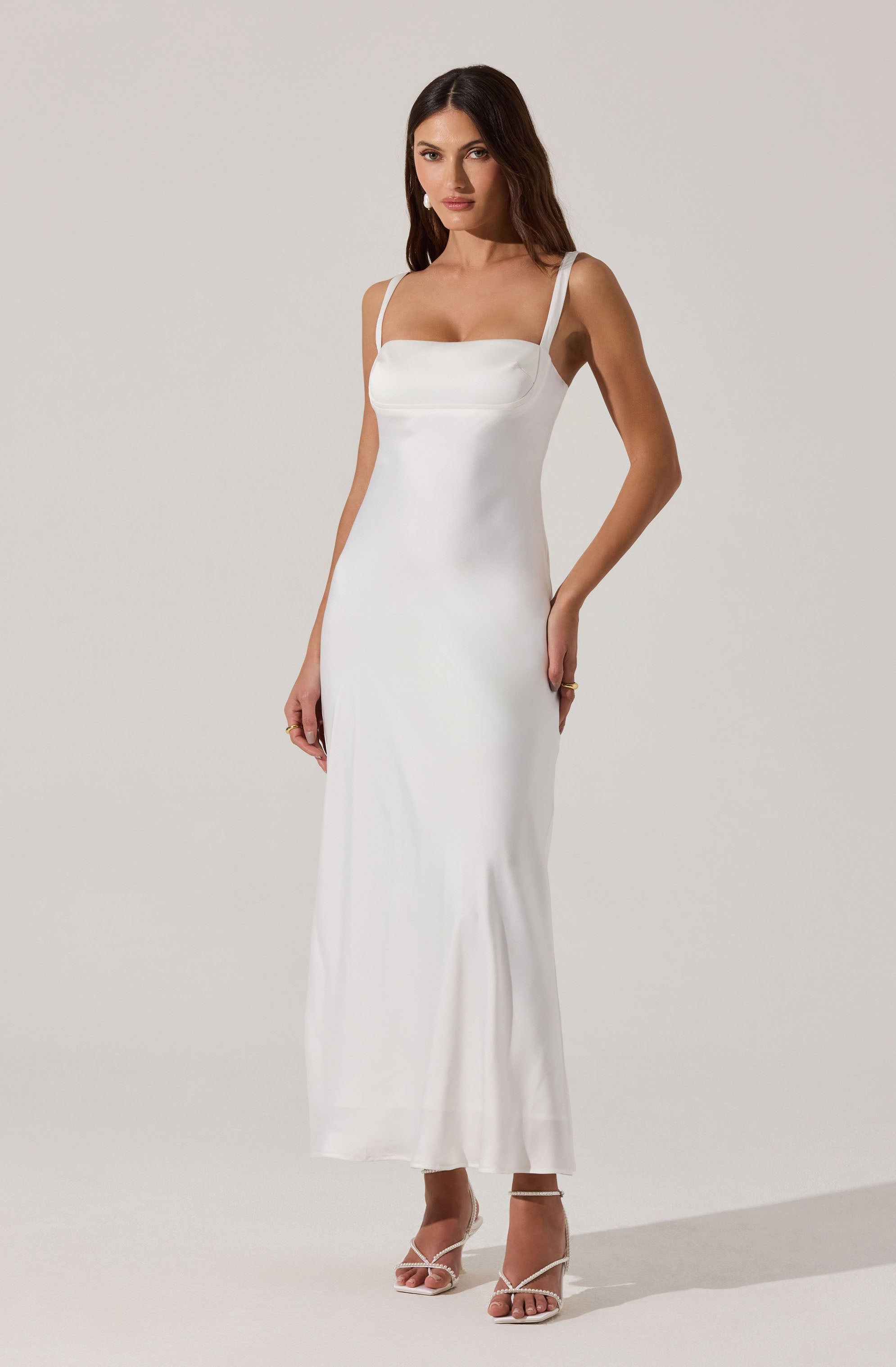 A woman is wearing a long, white, sleeveless dress with wide shoulder straps. The dress has a fitted bodice with a slight square neckline and a smooth, flowing skirt that reaches the floor. She is also wearing open-toe high heel sandals with thin straps.