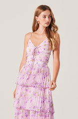 A woman is wearing a sleeveless, V-neck dress with thin spaghetti straps. The dress features a light, pastel-colored floral print in shades of pink, purple, and green. The top part of the dress is fitted, with a tie detail at the bust. The skirt has multiple tiered layers of fabric, with the top layer being slightly shorter and pleated, creating a flowing, layered look.