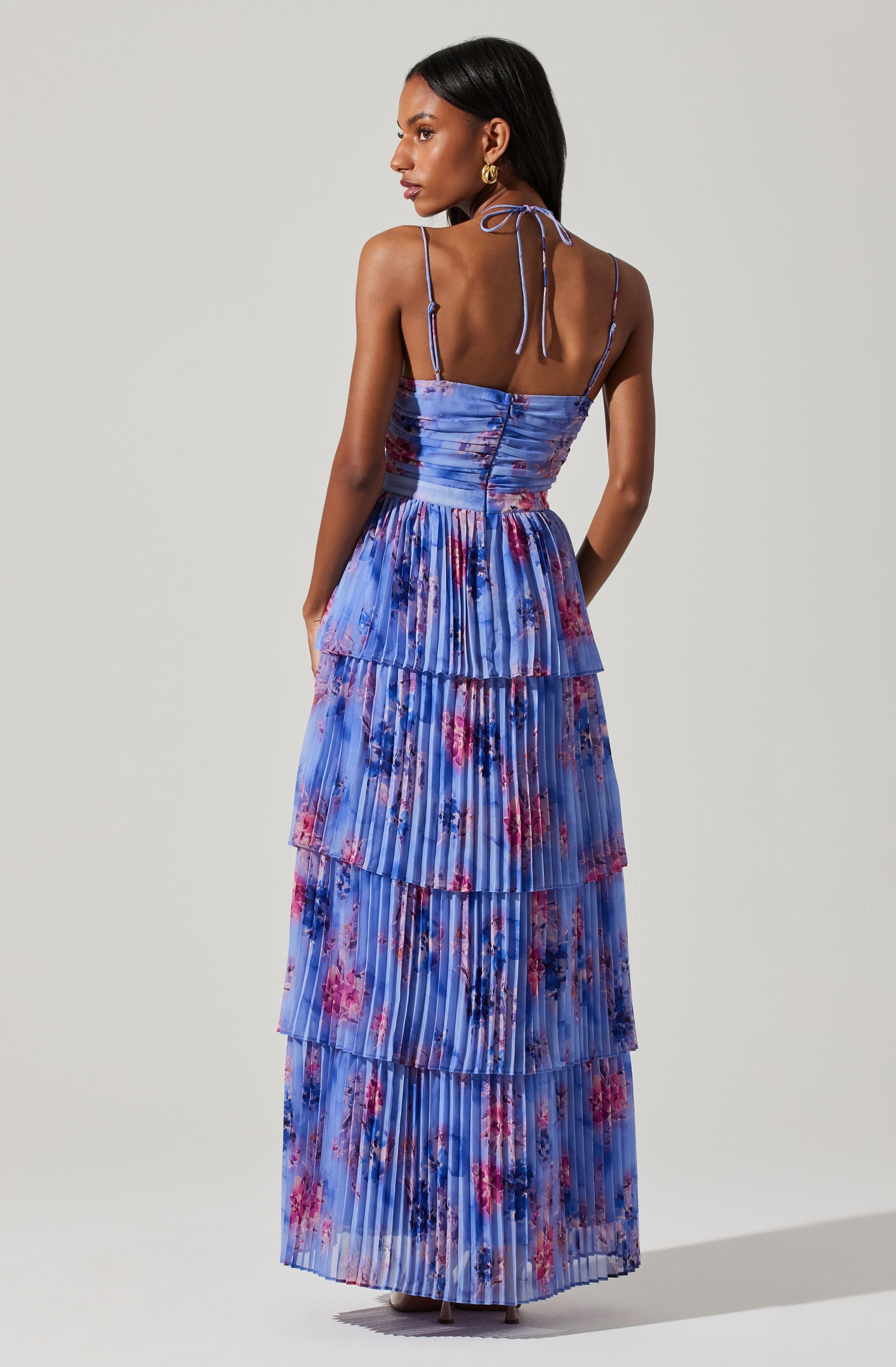 A woman is wearing a sleeveless, floor-length dress with thin, adjustable spaghetti straps tied in bows at the shoulders. The dress features a fitted bodice with a ruched design and a flowy, tiered skirt, all made from a pleated fabric with a floral pattern in shades of blue, pink, purple, and red.