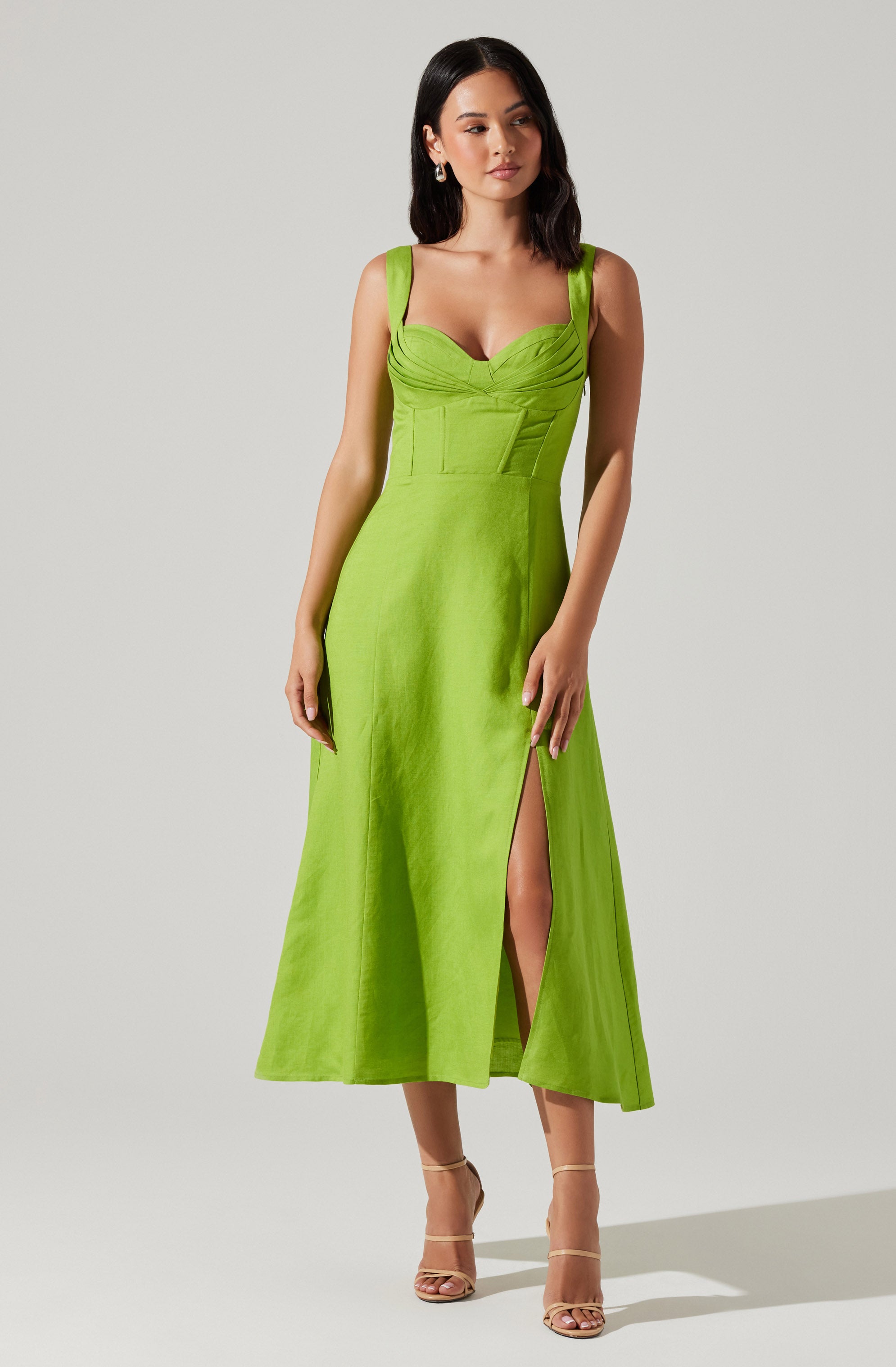 A woman is wearing a sleeveless, lime green dress with broad shoulder straps and a fitted bodice featuring ruching details. The dress has a high slit on one side, revealing her leg. She is also wearing beige high-heeled sandals with ankle straps.