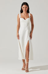 A woman is wearing a white sleeveless dress with wide straps, a fitted bodice, and a front slit that reveals her leg. She has paired the dress with beige high-heeled sandals that have ankle straps.