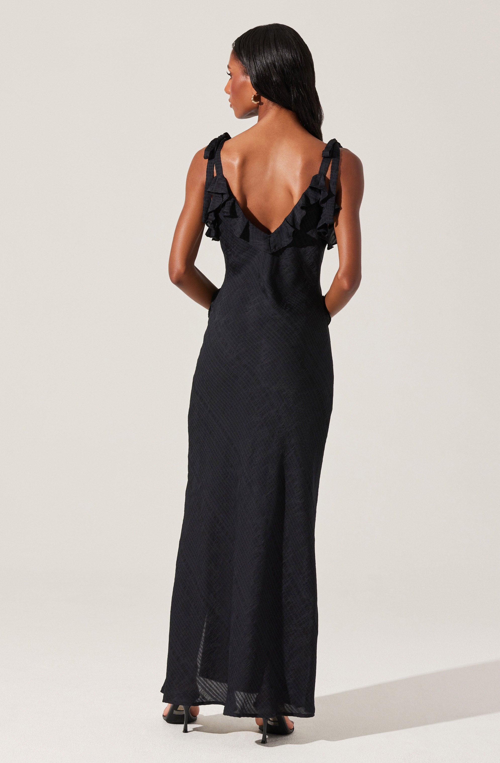 A woman is wearing a long, black sleeveless dress with textured fabric. The dress features ruffled shoulder straps that extend into a ruffled neckline, forming a V-shape in the back. She is also wearing black high-heeled sandals.
