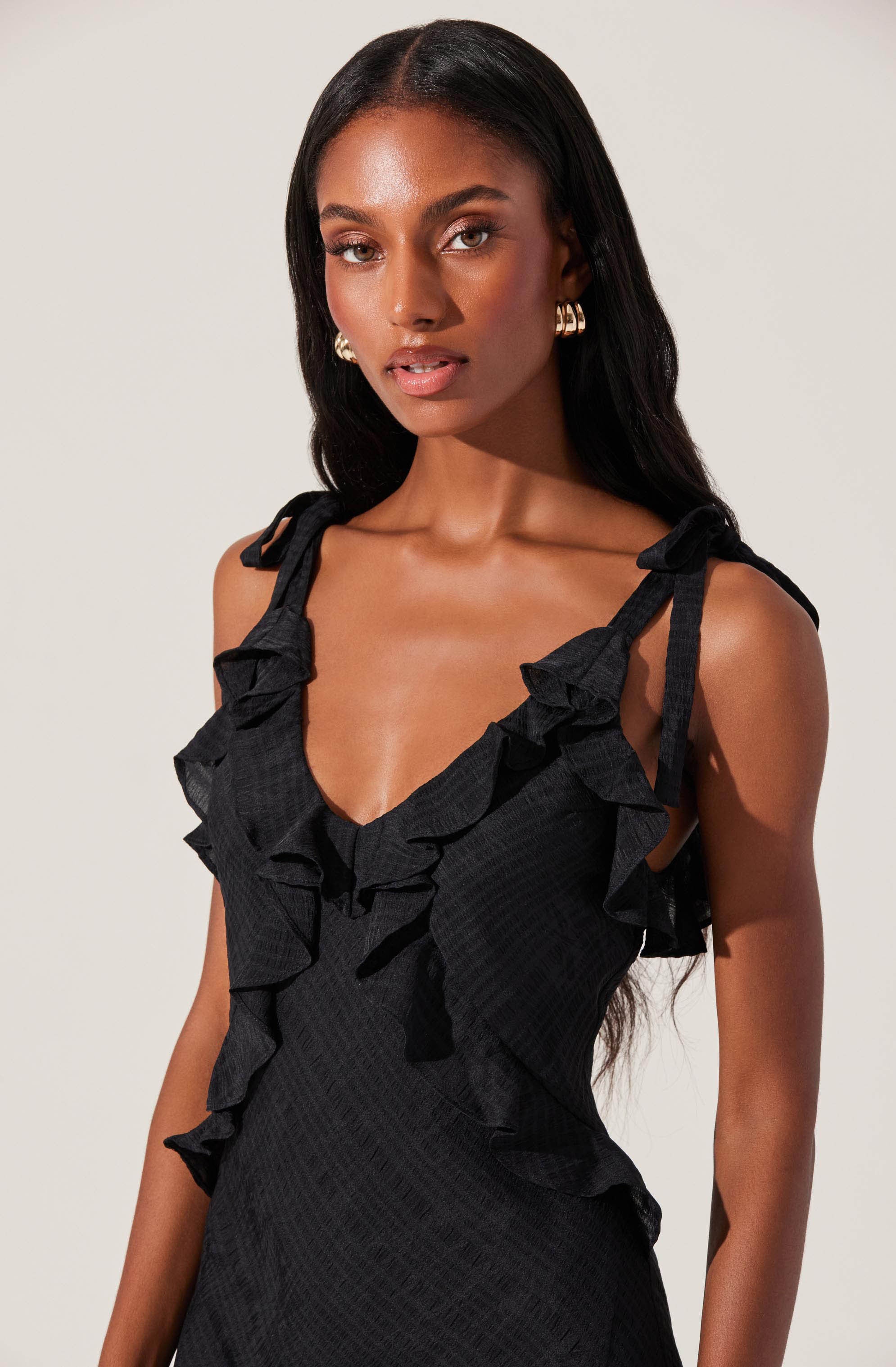 A woman is wearing a sleeveless black dress with ruffled straps and trim along the neckline and sides. The fabric has a subtle textured pattern.