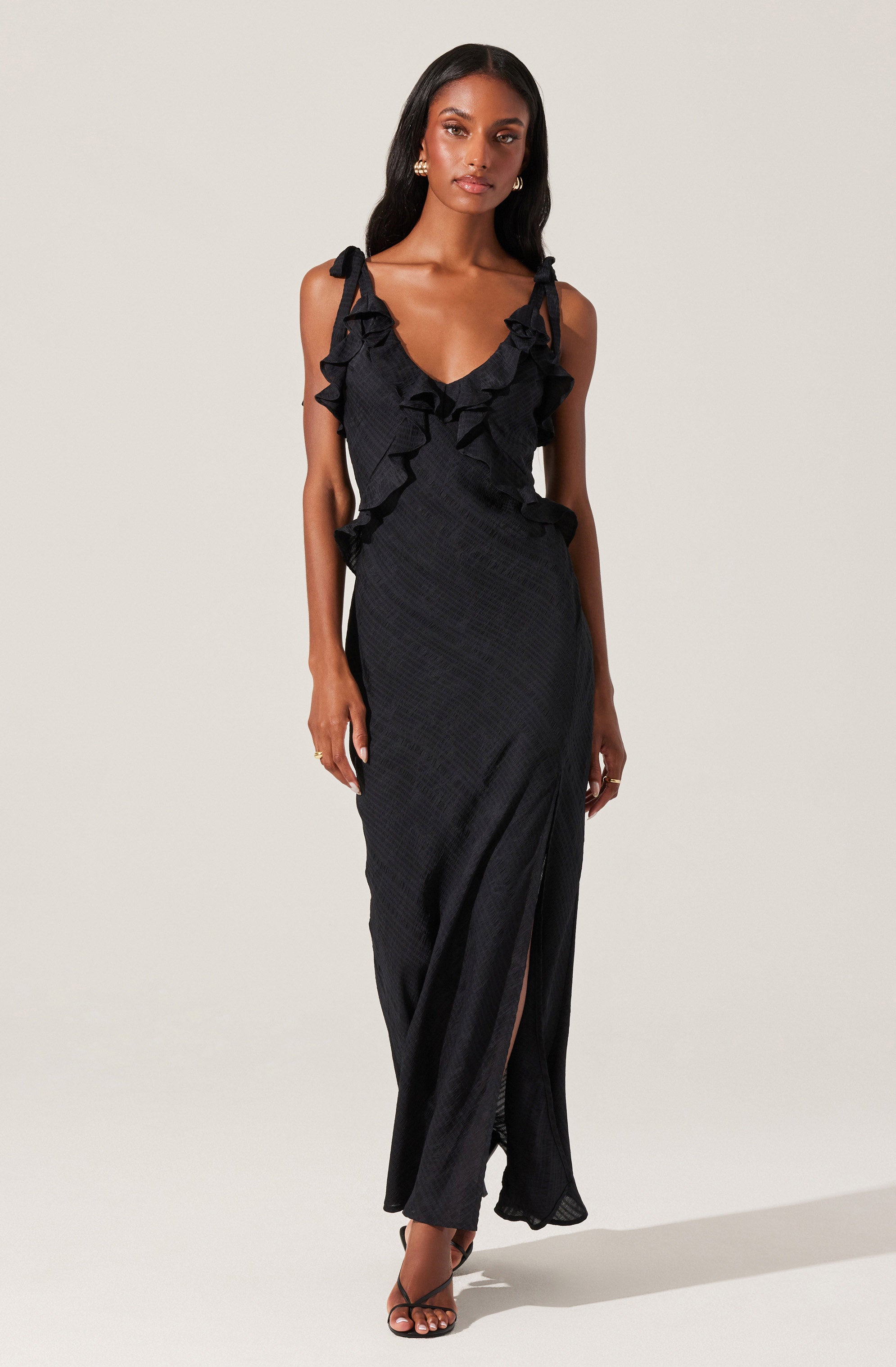 A woman is wearing a long, black dress with ruffled shoulder straps that extend around the neckline. The dress features a subtle textured pattern and a front slit that reveals part of her leg. She is also wearing a pair of open-toed black heels.
