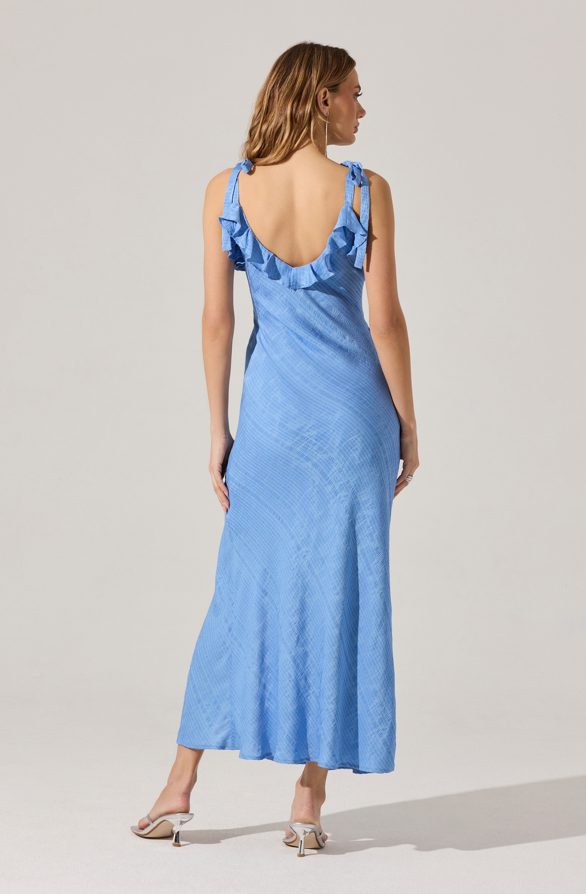 A woman is wearing a long, light blue dress with a subtle textured pattern. The dress features shoulder straps tied into bows and a ruffled neckline along the back. She is also wearing a pair of beige high-heeled shoes with ankle straps.