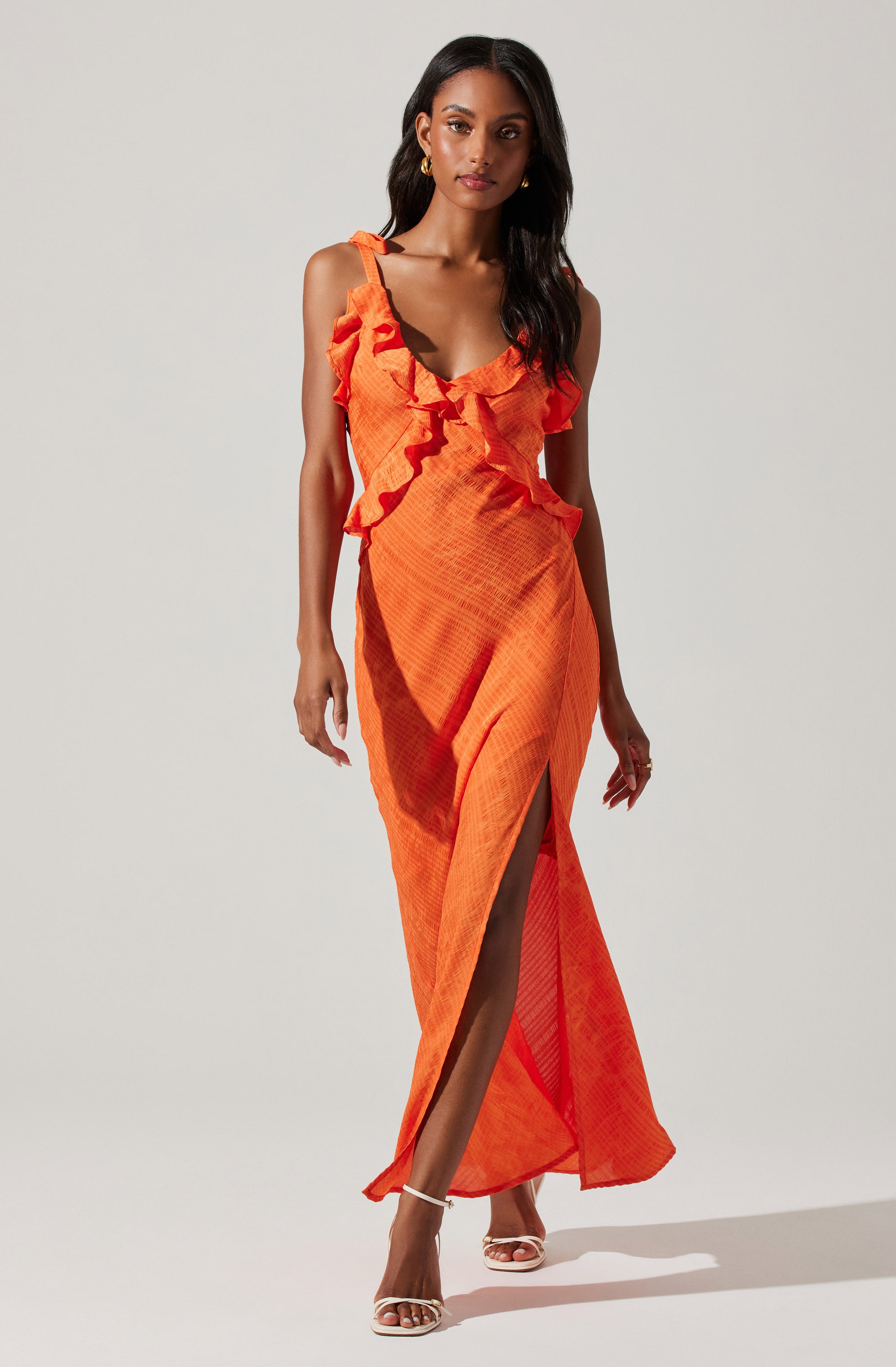 A woman is wearing an orange sleeveless maxi dress with ruffled straps and detailing around the neckline. The dress has a slit up the front, revealing her legs and high-heeled sandals. The fabric appears lightweight with a textured pattern.