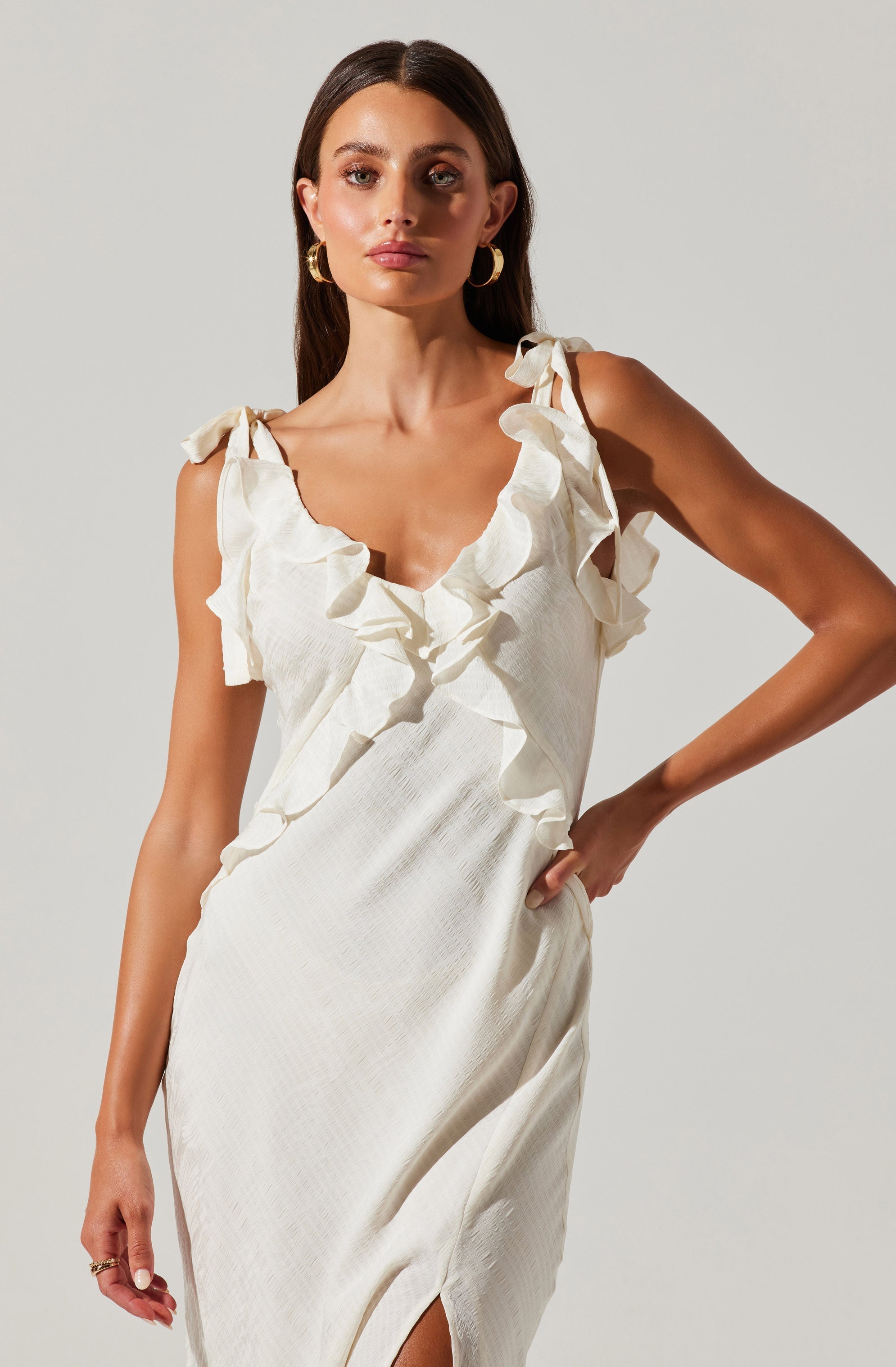 A woman wearing a sleeveless, cream-colored dress with ruffled shoulder straps and a ruffled neckline. The dress has a textured fabric and features a small slit at the front. She is accessorized with large gold hoop earrings and a ring on her left hand.