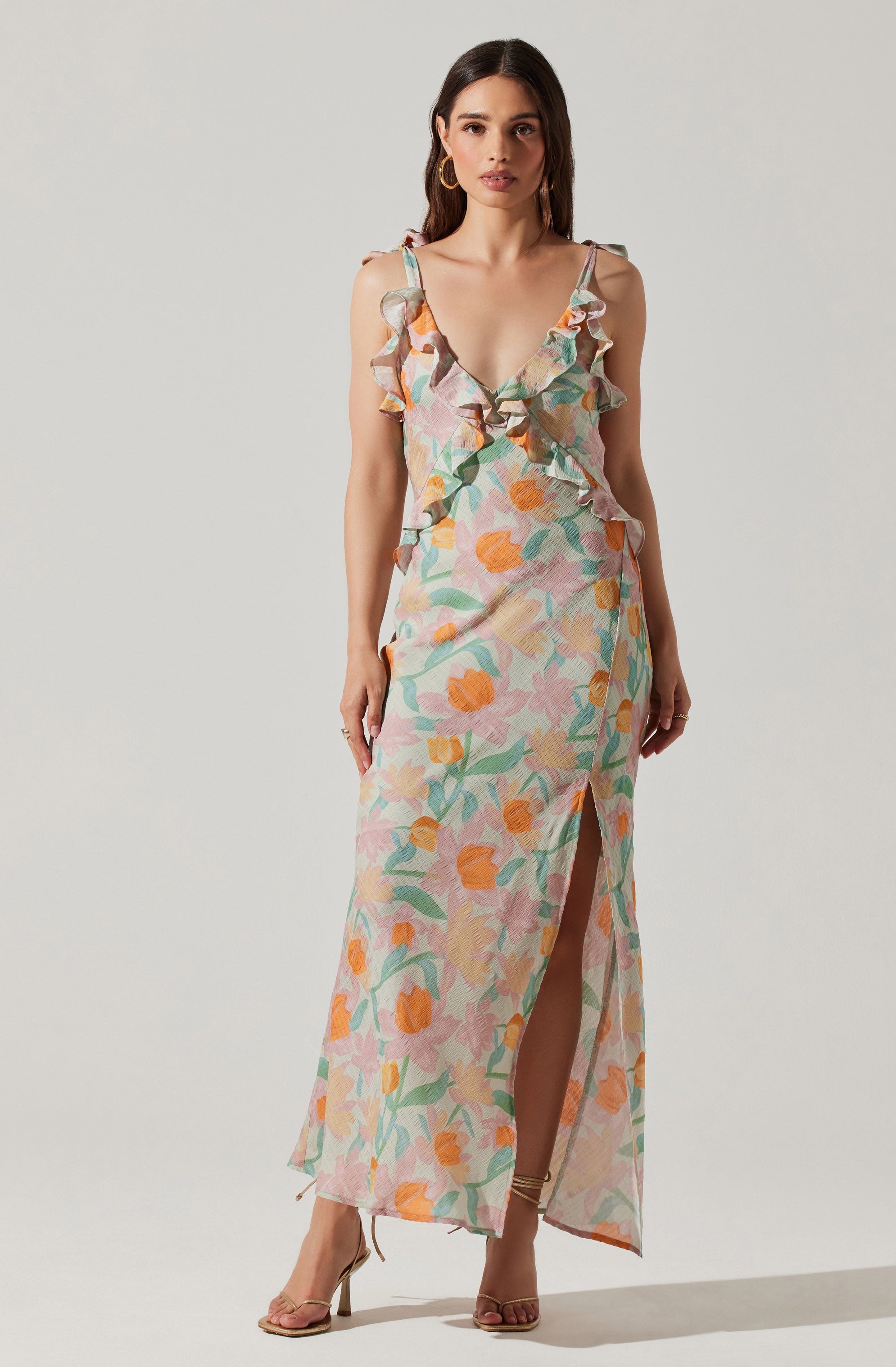 A woman is wearing a sleeveless, full-length dress with a floral print in pastel shades of pink, orange, green, and cream. The dress features ruffled shoulder straps, a deep V neckline, and a thigh-high slit in the front. She is also wearing high-heeled sandals and hoop earrings.