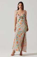 A woman is wearing a sleeveless, full-length dress with a floral print in pastel shades of pink, orange, green, and cream. The dress features ruffled shoulder straps, a deep V neckline, and a thigh-high slit in the front. She is also wearing high-heeled sandals and hoop earrings.