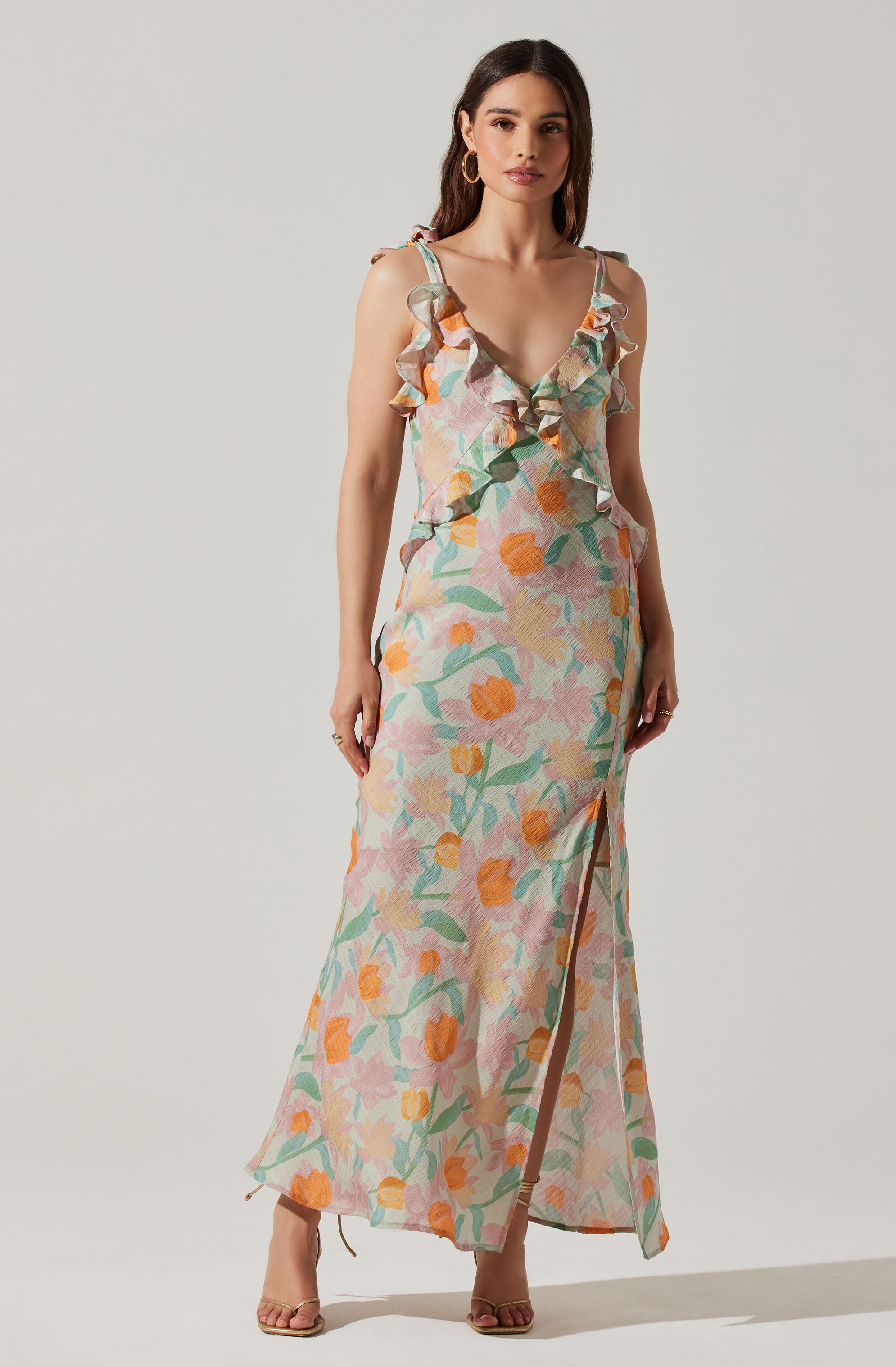 A woman is wearing a long, flowy dress with a colorful floral pattern featuring shades of pink, orange, green, and beige. The dress has ruffled shoulder straps and a plunging V-neckline. It also has a high slit on one side, revealing part of her leg. She is accessorized with large, circular hoop earrings and neutral-colored high-heeled sandals.
