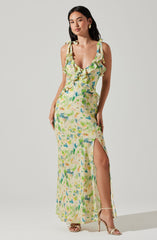 A woman is wearing a long, sleeveless dress with shoulder ties and a ruffled neckline. The dress features a colorful, abstract floral pattern in shades of yellow, green, blue, and pink. It has a high slit on one side, revealing her leg. She is also wearing gold hoop earrings and beige high-heeled sandals.