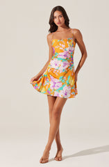 A young woman is wearing a sleeveless, floral print dress with thin spaghetti straps. The dress features a vibrant pattern of large pink, purple, yellow, and green flowers on an orange background. She is also wearing open-toe, nude-colored high-heeled sandals.