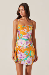 A woman is wearing a sleeveless dress with thin spaghetti straps. The dress features a bright orange background with a large floral pattern in shades of purple, pink, yellow, and teal. The dress has a fitted bodice and a slightly gathered detail on one side, creating a subtle drape effect.