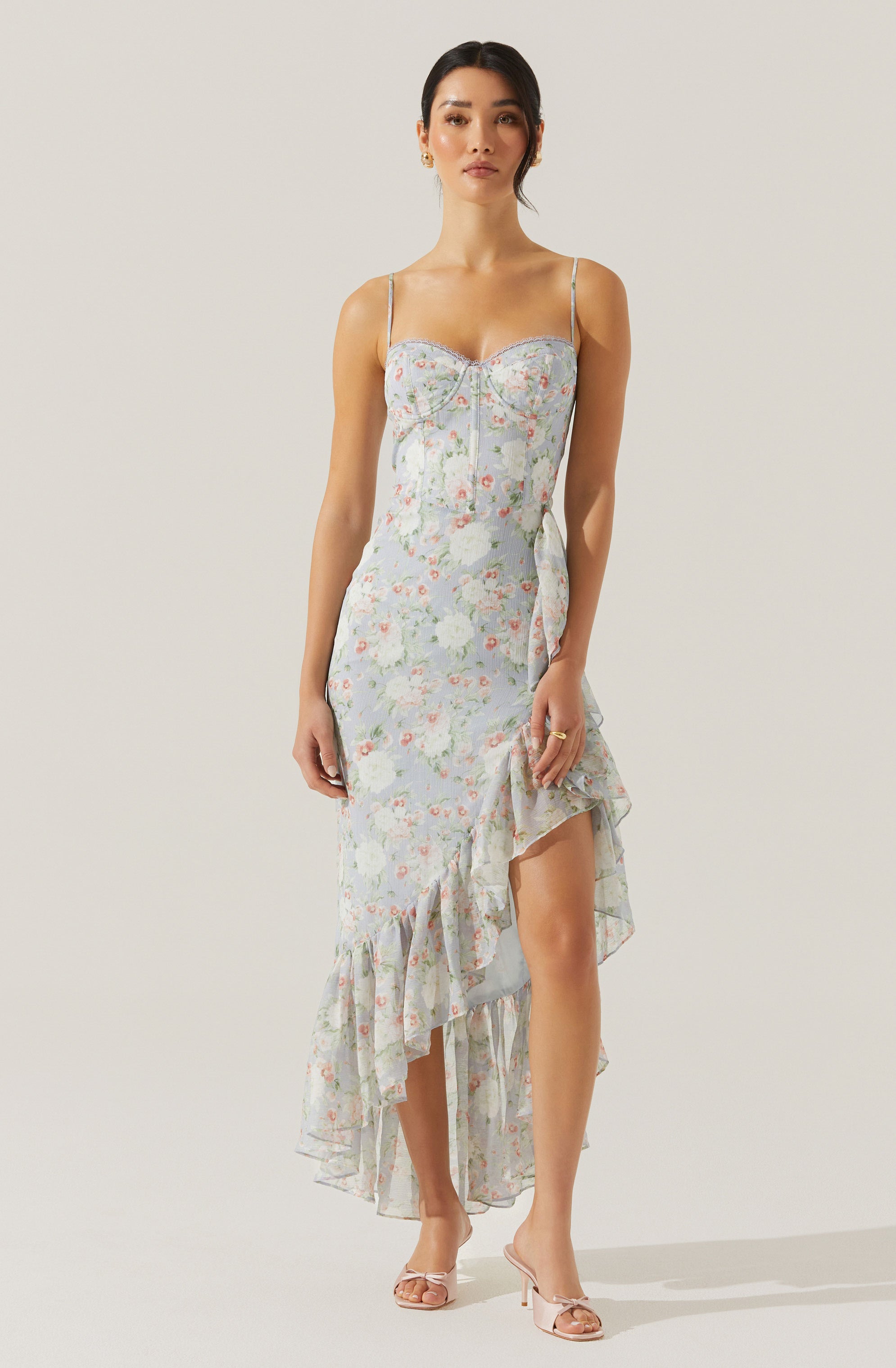 A woman is wearing a sleeveless, fitted dress with thin spaghetti straps. The dress has a light, floral pattern with soft pastel colors and features a ruffled hemline that is asymmetrical, longer in the back and shorter in the front. The fabric appears lightweight and flowy, and the dress has a subtle zipper detail at the bust area. She is also wearing nude high-heeled sandals.