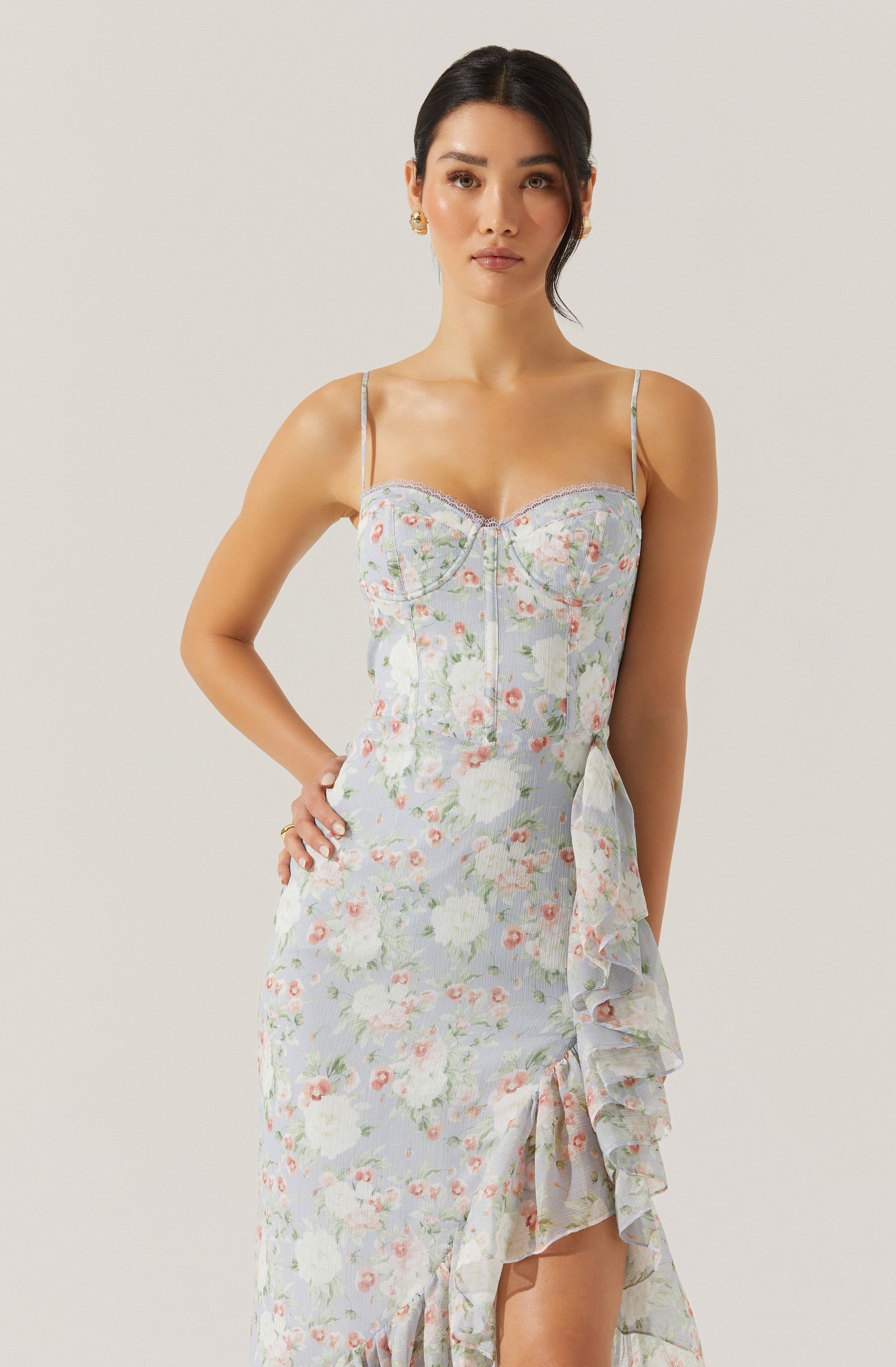 A woman is wearing a sleeveless, light-colored dress with thin spaghetti straps. The dress features a floral pattern with pastel shades of white, pink, green, and blue. It has a fitted bodice with a visible zipper front, and the skirt has a ruffled, asymmetrical design that drapes down the side.