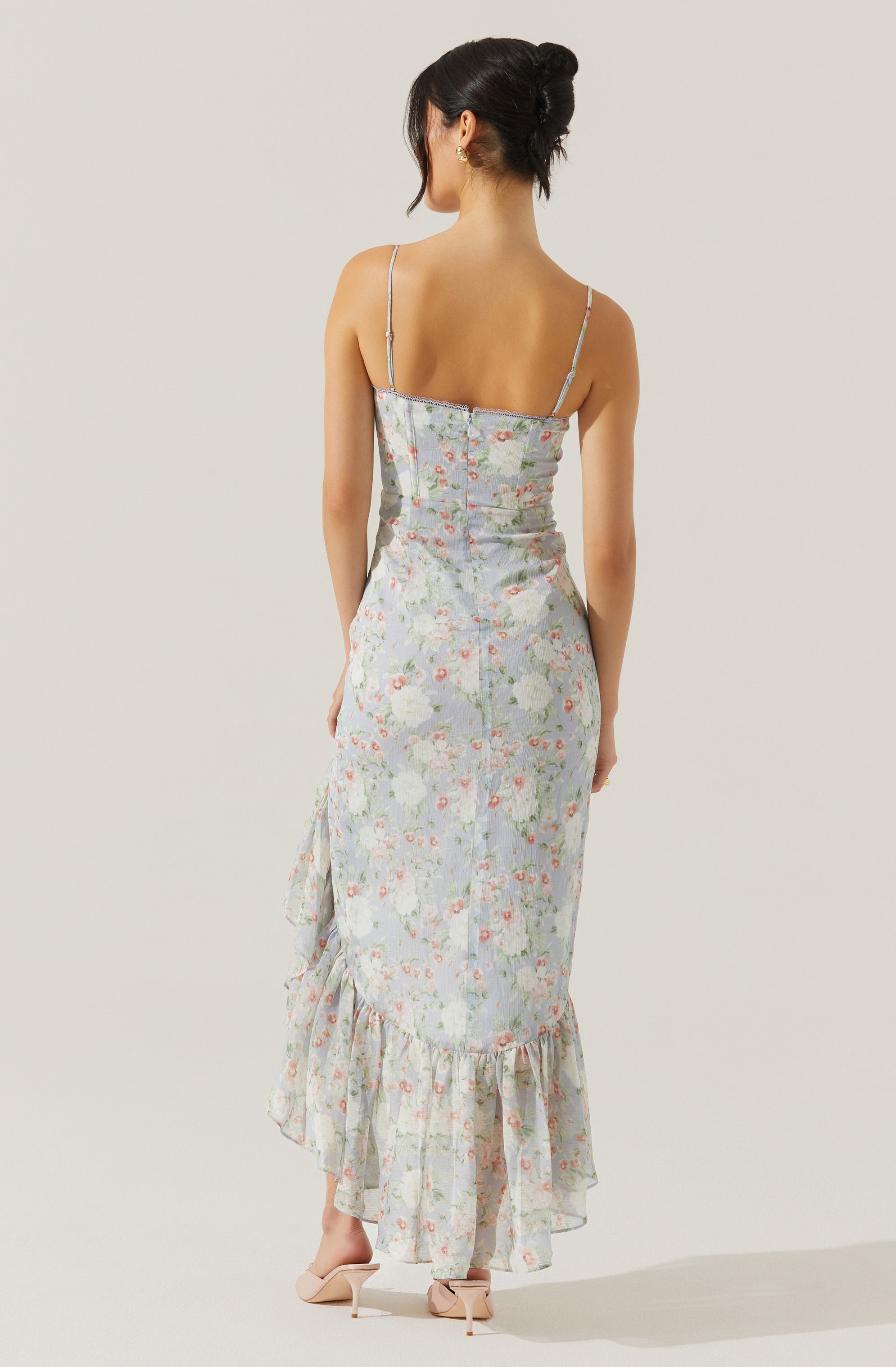 A woman is wearing a light-colored, floral-patterned dress with a fitted bodice and thin spaghetti straps. The dress has a ruffled hem that falls to ankle length and features a delicate, pastel floral print. She is also wearing nude high-heeled sandals.
