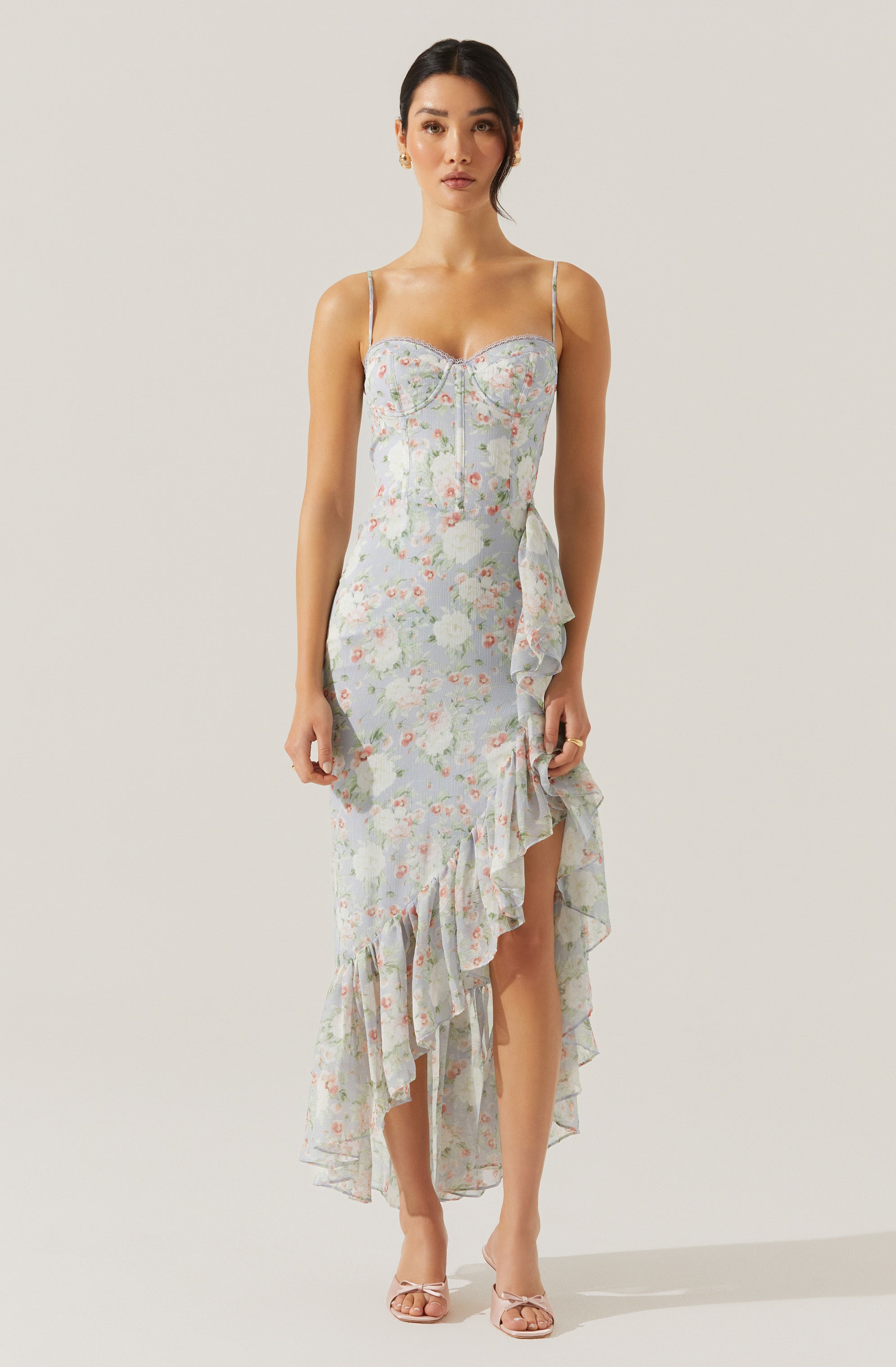 A woman is wearing a light-colored, floral-patterned dress with thin spaghetti straps. The dress features a fitted bodice with a sweetheart neckline and a ruffled, asymmetrical hemline that cascades down the front and sides, creating a high-low effect.