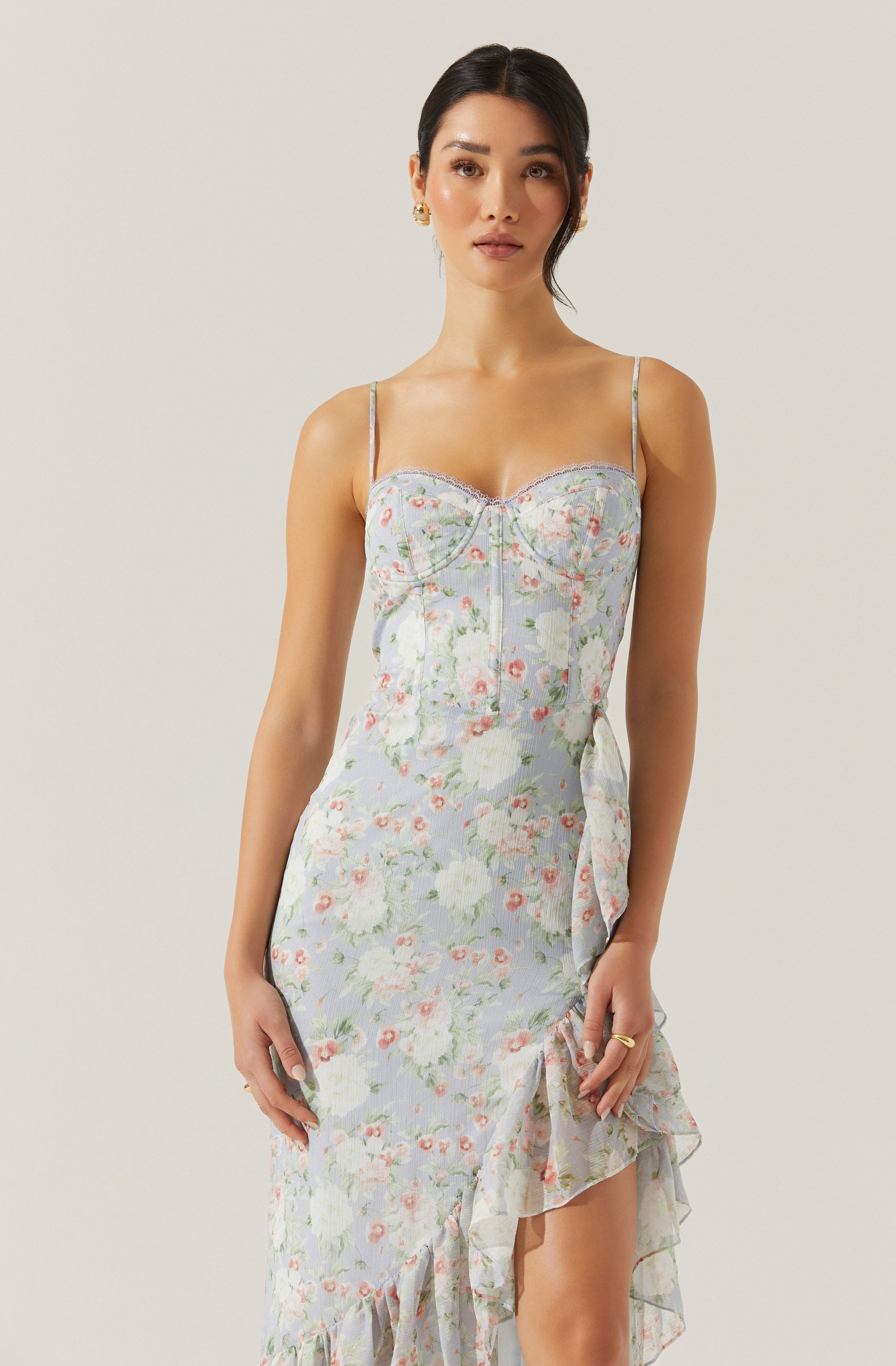 A woman is wearing a sleeveless, fitted dress with thin spaghetti straps. The dress has a light, pastel-colored floral pattern with shades of pink, white, and green. It features a ruffled hemline that cascades down the side, adding a decorative touch. The fabric appears soft and lightweight, suitable for warm weather.