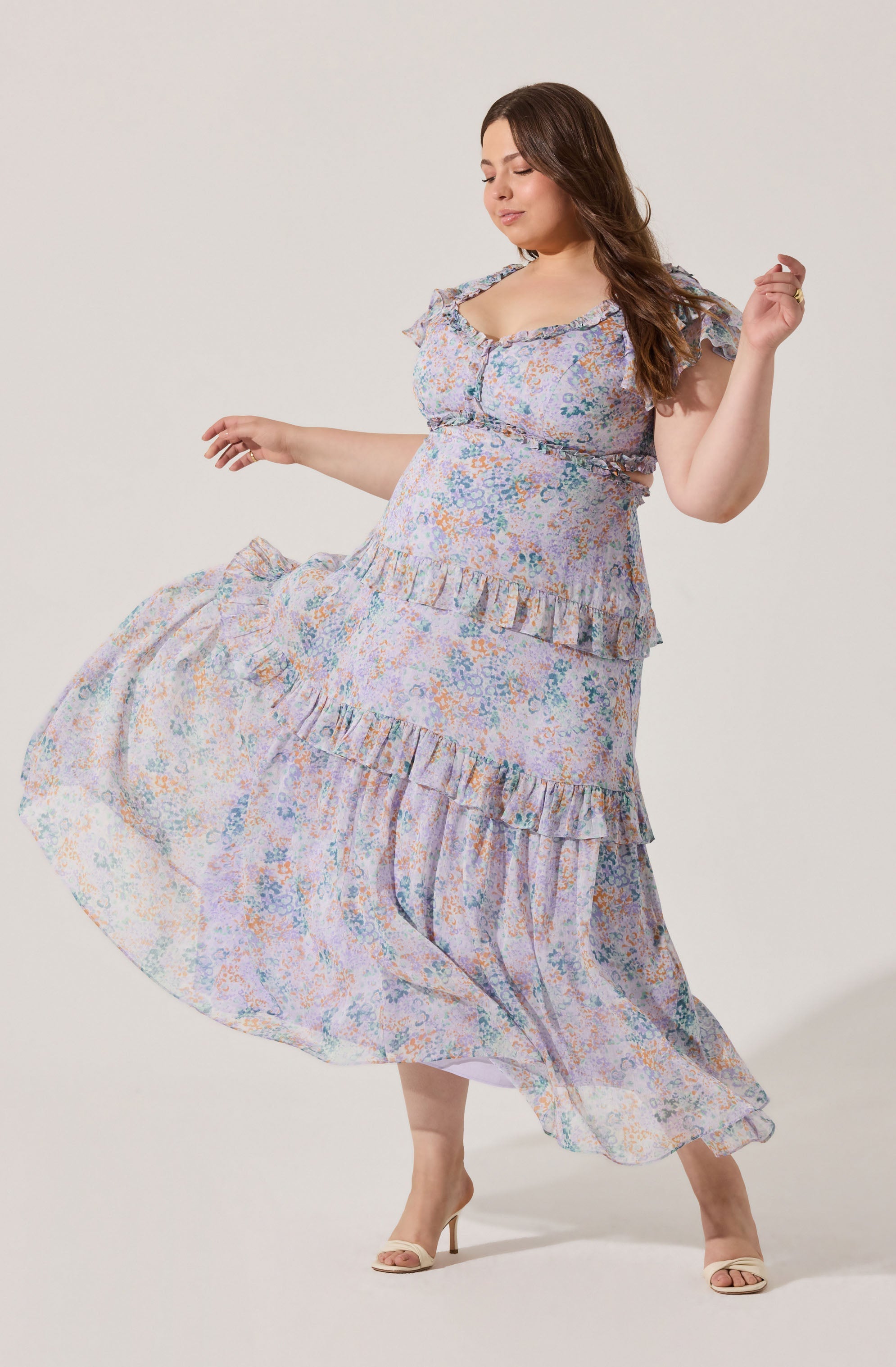 A woman is wearing a light-colored, floral-patterned dress with ruffled details along the neckline, sleeves, and around the waist. The dress has a flowing, layered skirt and appears to be made of a lightweight, airy fabric. She is wearing nude, open-toe heels.