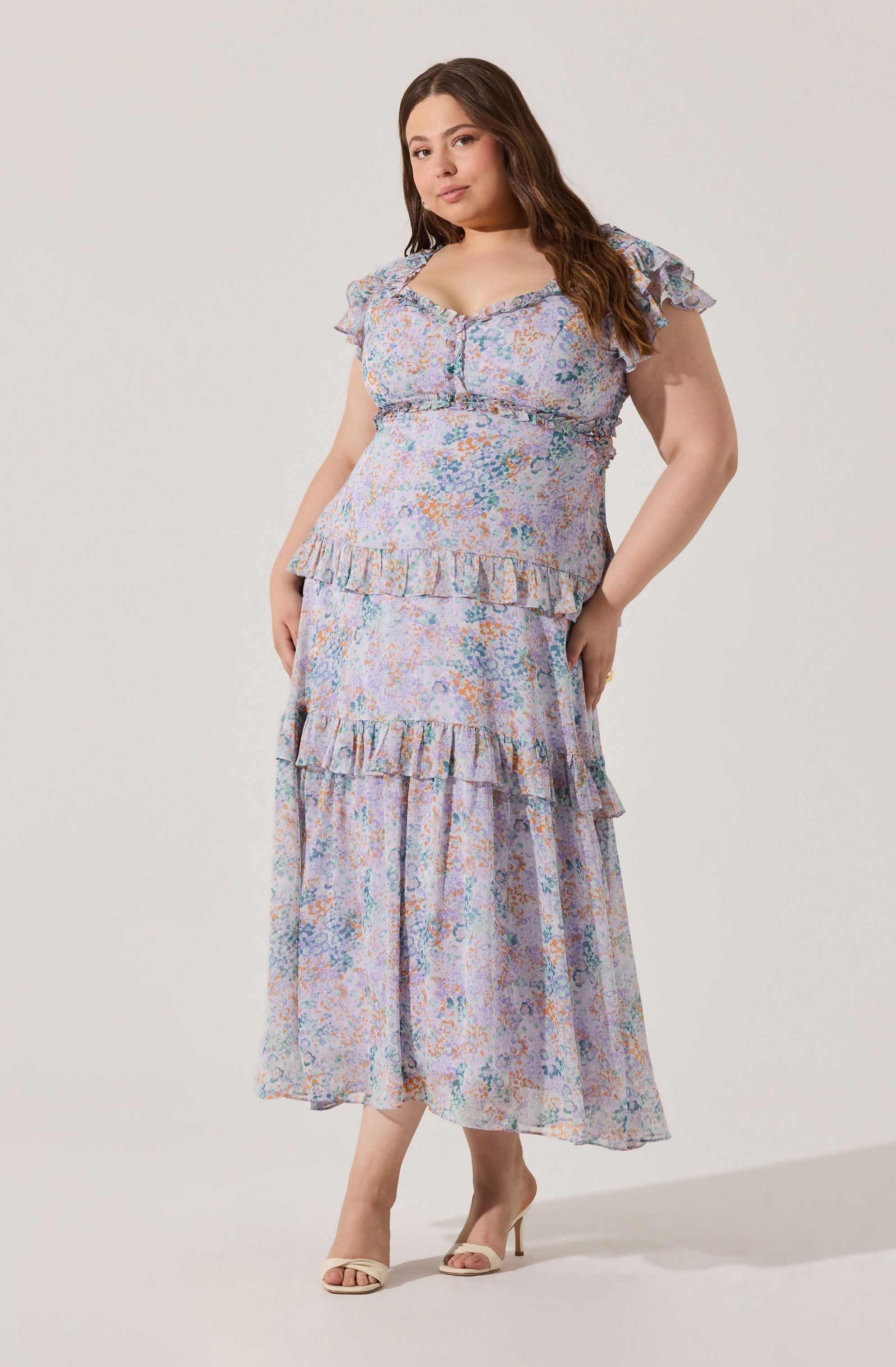 A woman is wearing a light, floral-patterned dress with a pastel color palette that includes shades of blue, pink, and orange. The dress has a square neckline with small ruffle details along the edges, short ruffled sleeves, and a fitted bodice. It features two tiers of ruffles, one around the waist and another near the hem of the skirt, which adds volume and texture. She is also wearing open-toed, high-heeled sandals in a light or neutral color.