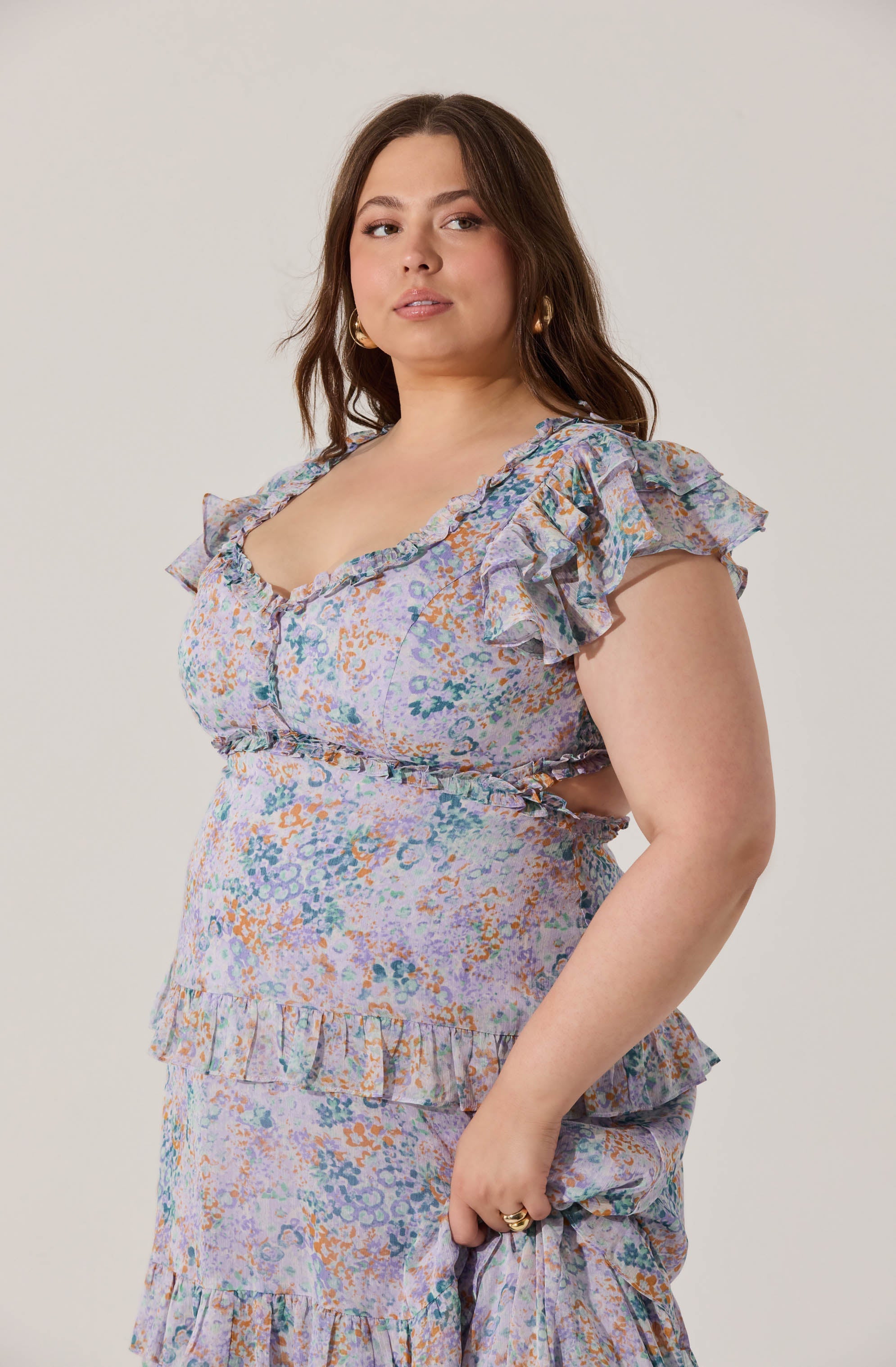 A woman is wearing a matching two-piece outfit with a pastel-colored floral pattern. The top features ruffled cap sleeves and a sweetheart neckline, with ruffles along the edges. The skirt has a high waist, a ruffle detail near the bottom, and a tiered design with additional ruffles.