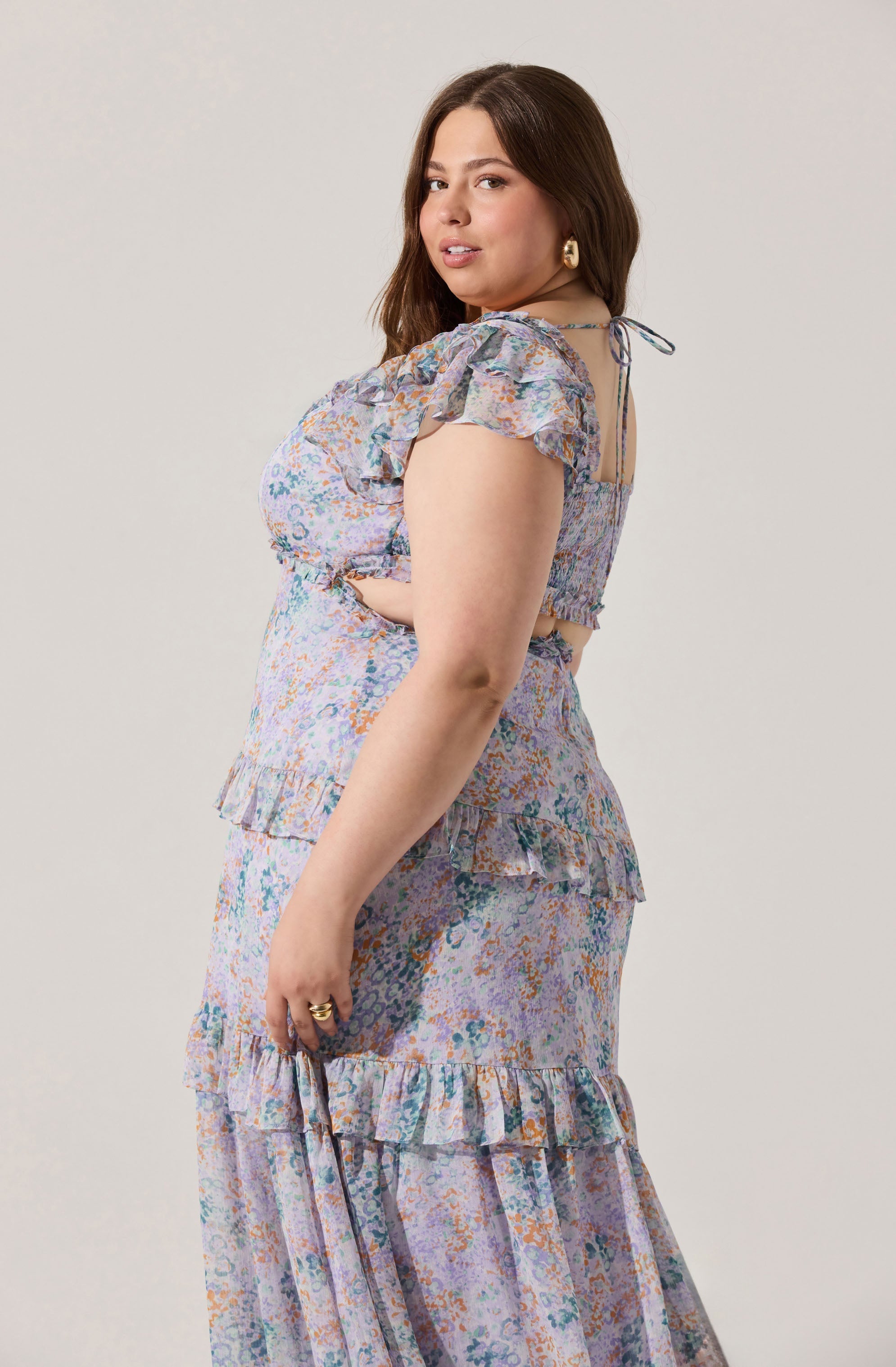 A woman is wearing a matching two-piece outfit with a pastel-colored abstract floral print. The top has short, ruffled sleeves with bows tied at the shoulders and a smocked bodice. The skirt is long with tiered ruffles at the hem.