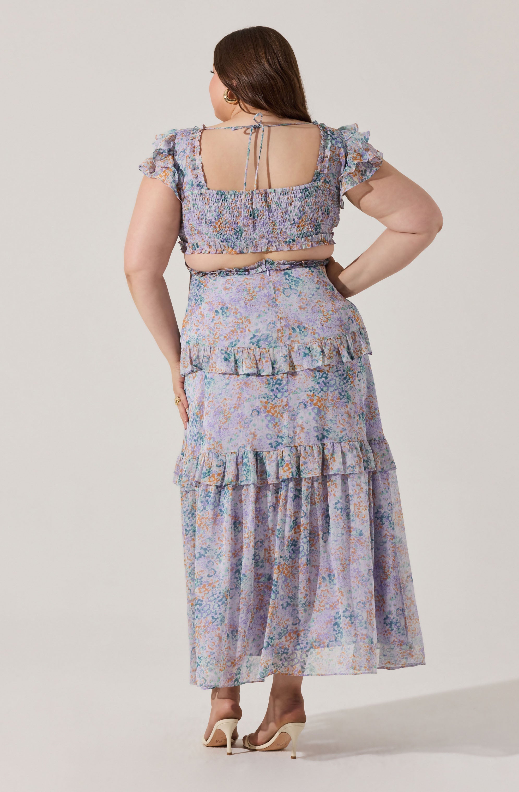 A woman is wearing a matching two-piece outfit consisting of a crop top and a long skirt. The crop top has short, ruffled sleeves, an elasticized, smocked bodice, and a square neckline with thin ties at the back. The high-waisted skirt features a tiered, ruffled design with a flowing, sheer fabric. The fabric has a multicolored floral pattern with shades of blue, purple, orange, and green. She is also wearing nude-colored high-heeled shoes.