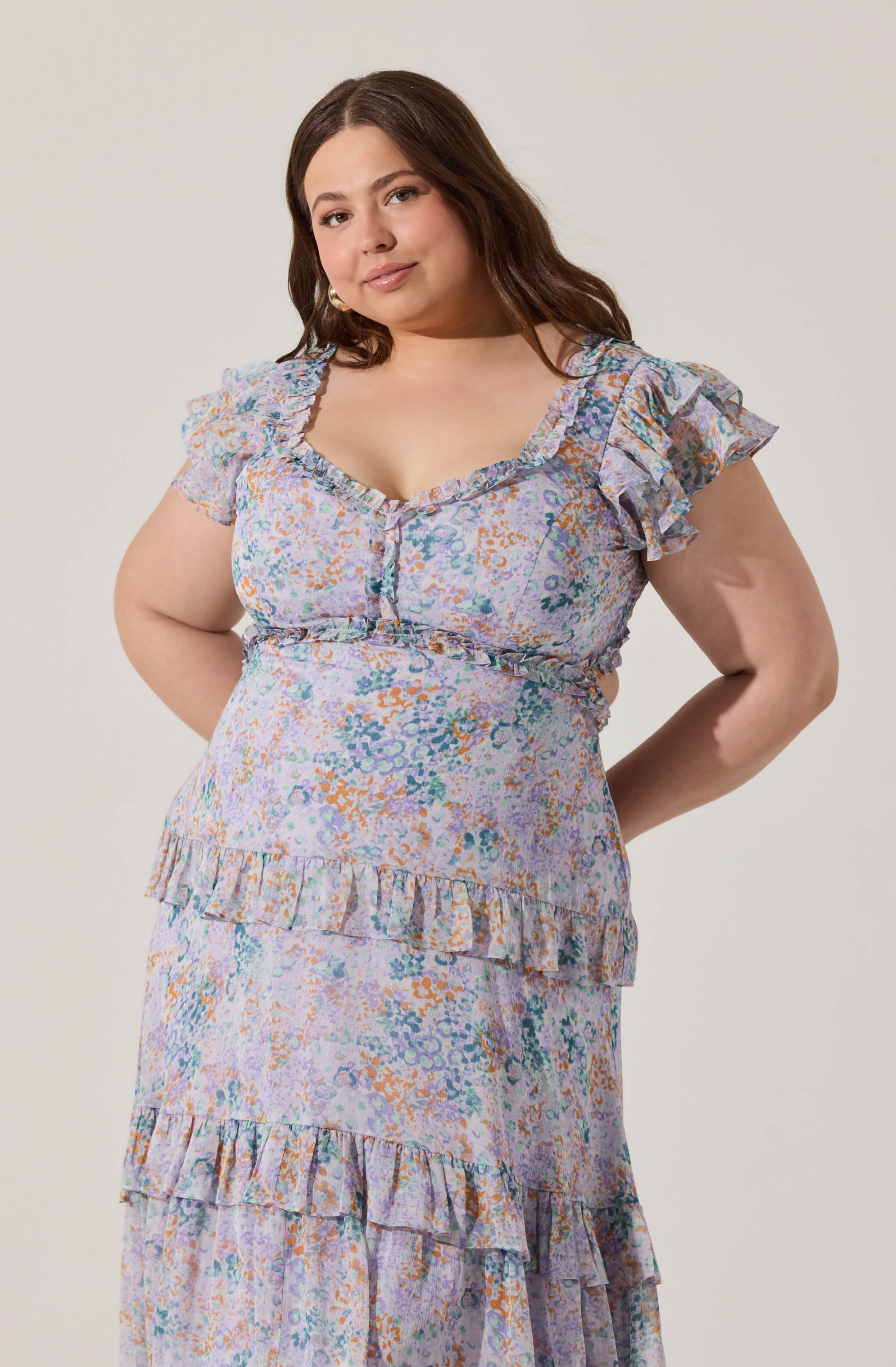 A woman is wearing a light-colored dress with a floral pattern featuring shades of purple, orange, blue, and green. The dress has short, ruffled sleeves with a frilled trim along the neckline. It includes multiple tiers with ruffle accents, creating a layered look. The fabric appears lightweight and airy.
